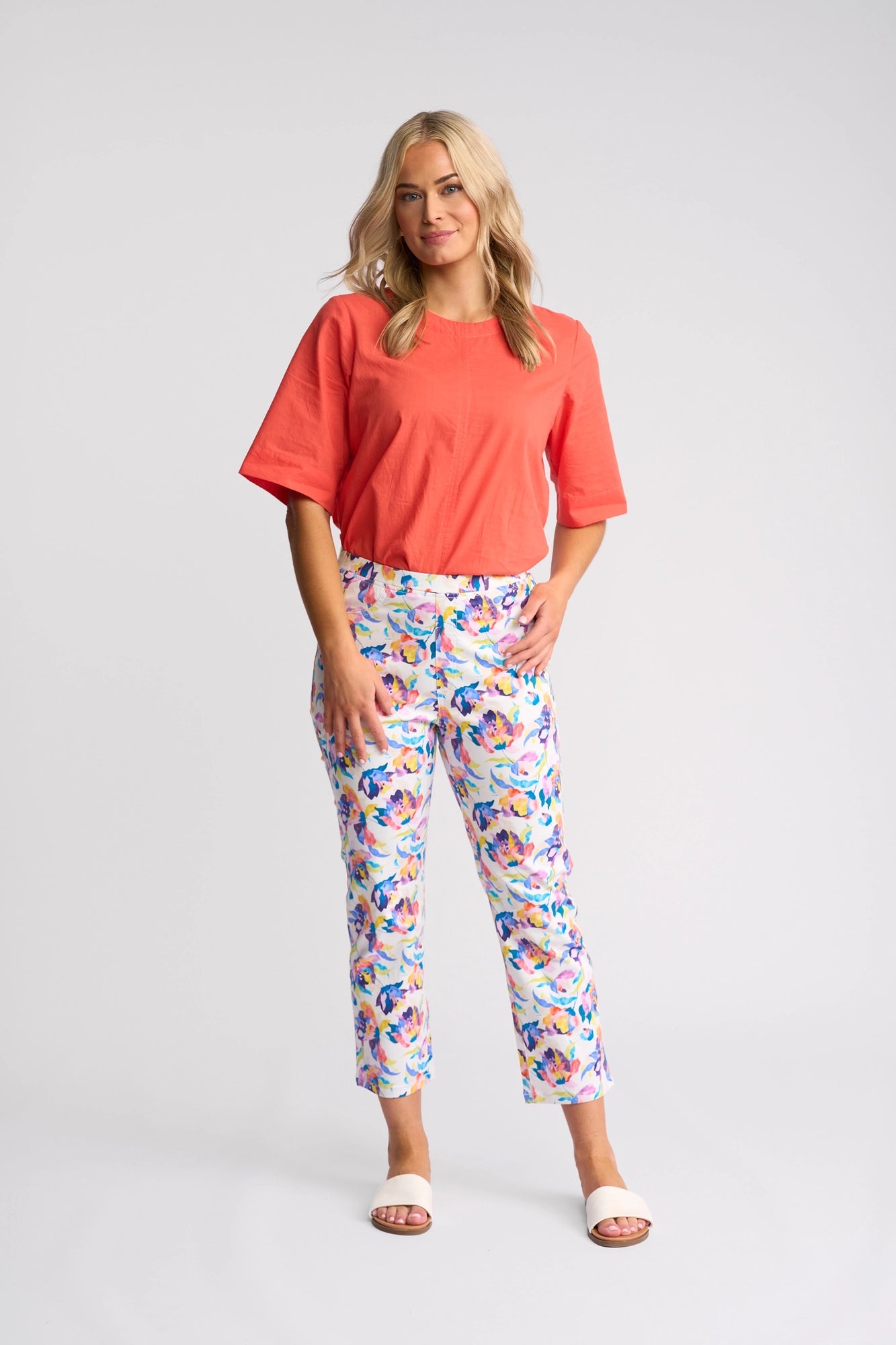 A woman stands in a studio with her left hand in her pocket and a slight smile, wearing a coral top, Vassalli Sierra 7/8 Pant - Demi in a floral print crafted from stretch cotton, and white slide sandals.