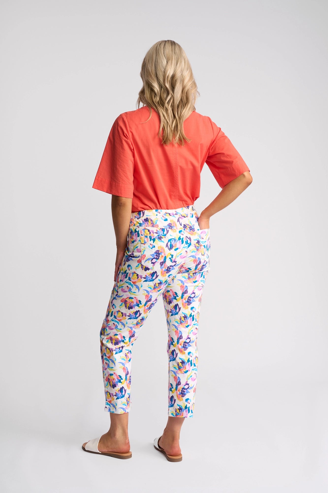 A woman with blonde hair, seen from behind, wears a coral short-sleeve top, white slide sandals, and the Vassalli Sierra 7/8 Pant - Demi: pull-on slim-leg floral print pants crafted in stretch cotton. She stands against a plain white background.