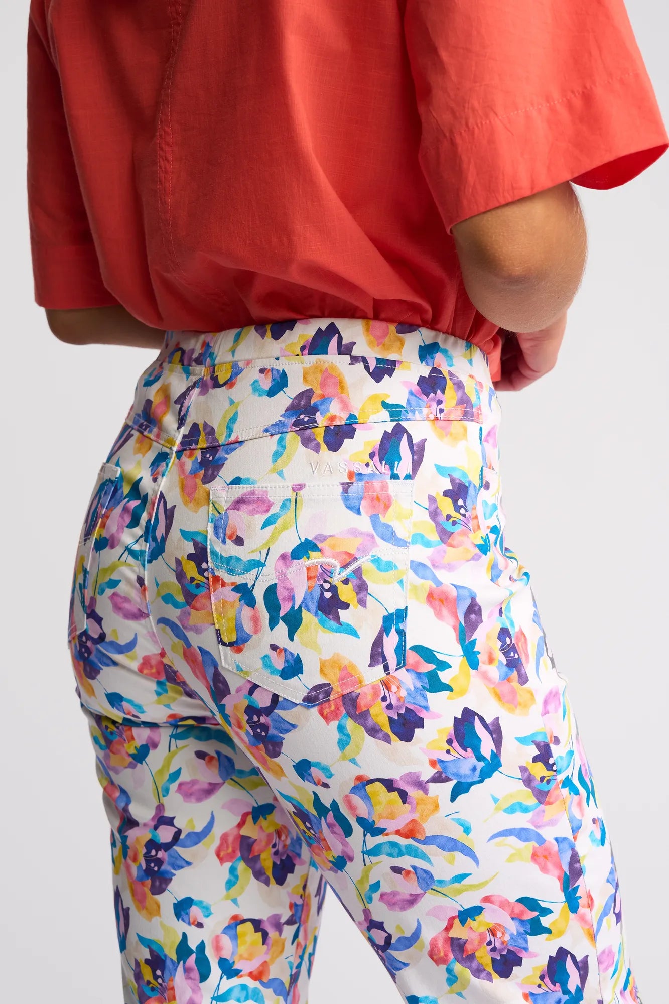 A person shown from the back, standing against a plain light background, is wearing a short-sleeved red-orange shirt and Vassalli’s Sierra 7/8 Pant - Demi: white pull-on slim-leg pants featuring a colorful floral print.