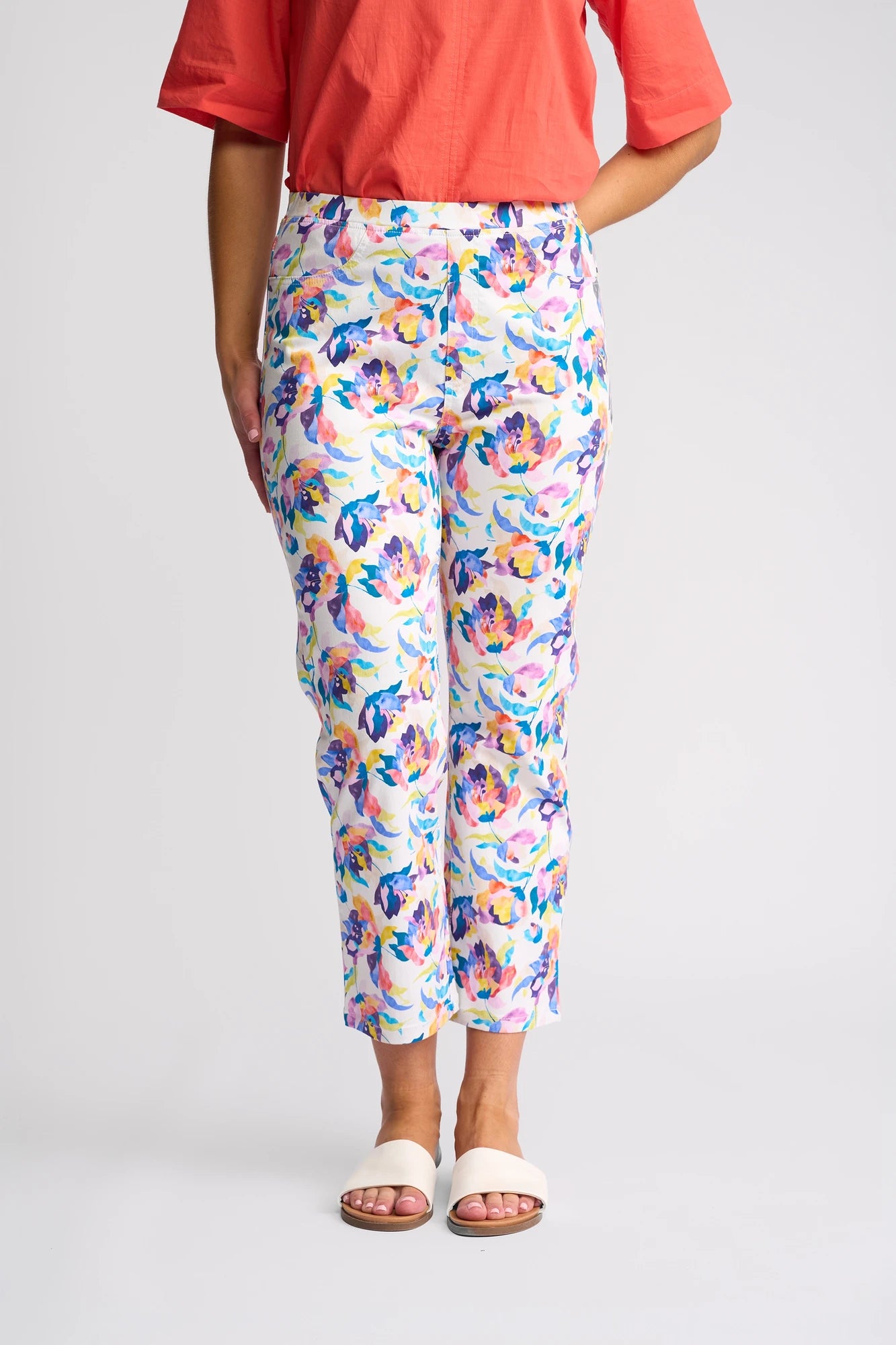 A person is wearing a coral short-sleeve top, Vassalli Sierra 7/8 Pant - Demi pull-on slim-leg floral print pants crafted from stretch cotton, and white sandals, standing against a plain white background.