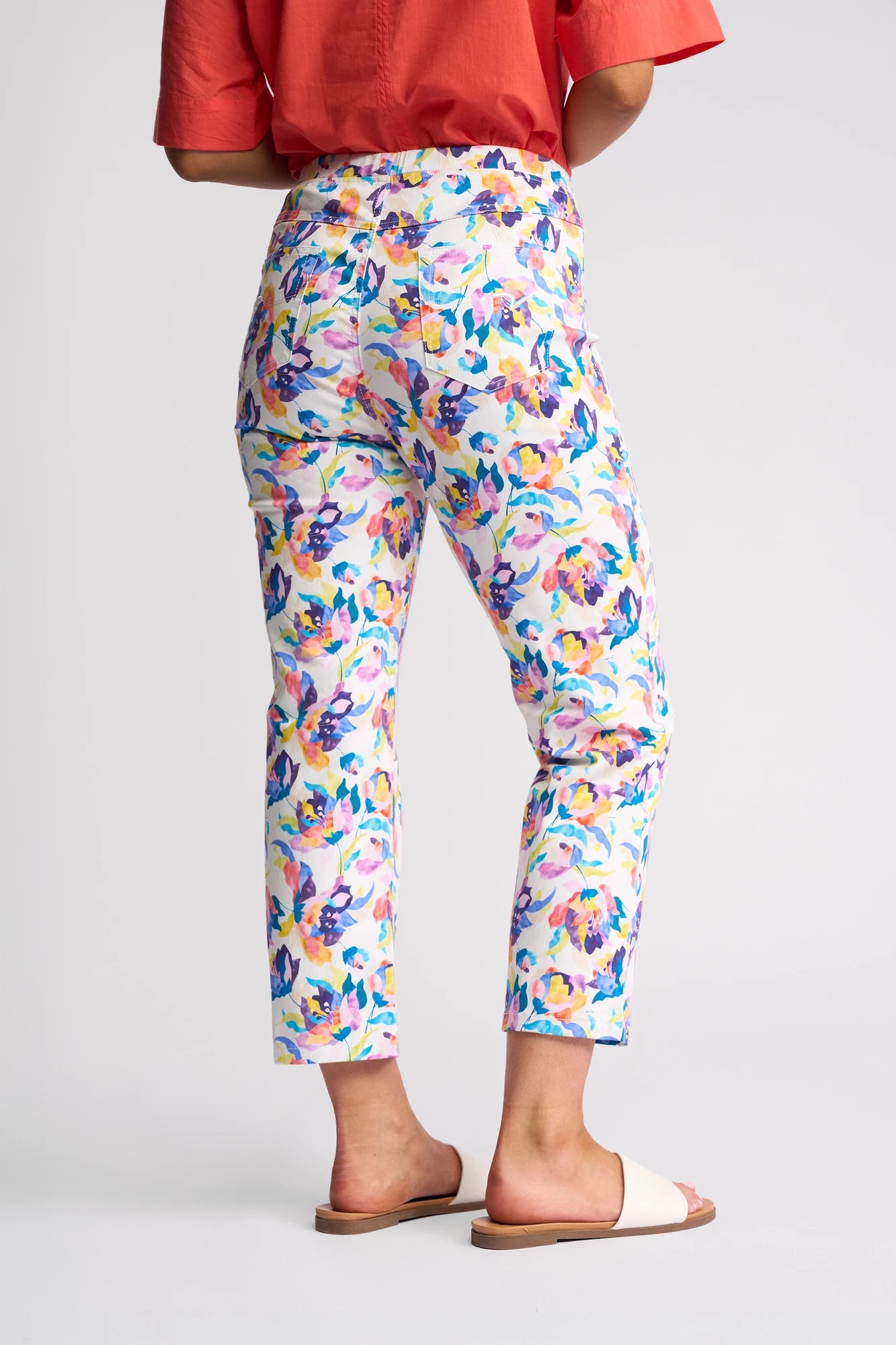 A person wearing Vassalli’s Sierra 7/8 Pant in the Demi floral print, paired with a red short-sleeve top and white slide sandals, stands against a plain light background, seen from the back.