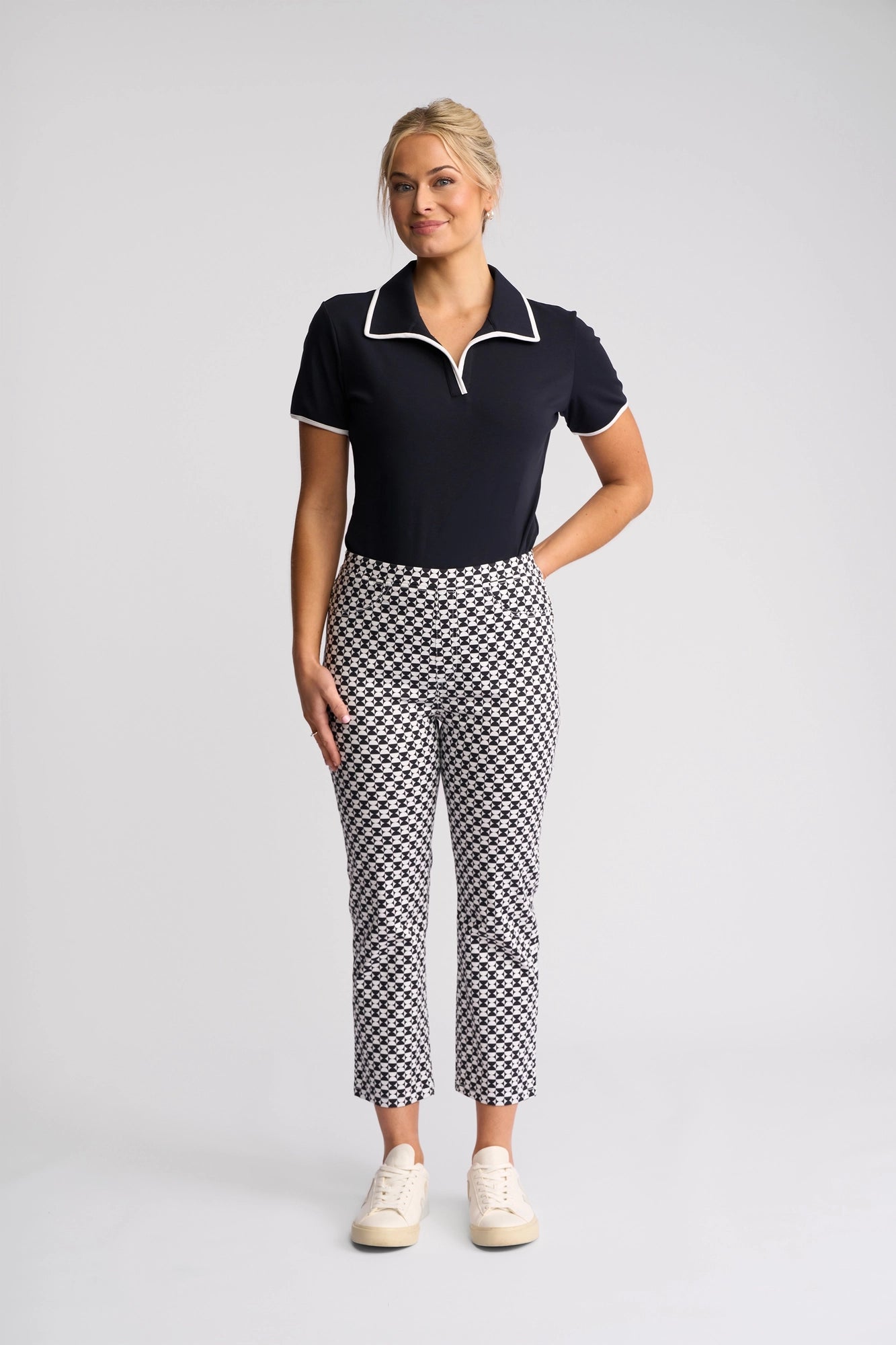 A woman stands against a plain background, wearing a navy collared polo shirt, Vassalli Sierra 7/8 Pant – Dixie in a black-and-white print crafted from stretch cotton, and white sneakers. She has blonde hair styled up and is smiling softly with one hand behind her back.