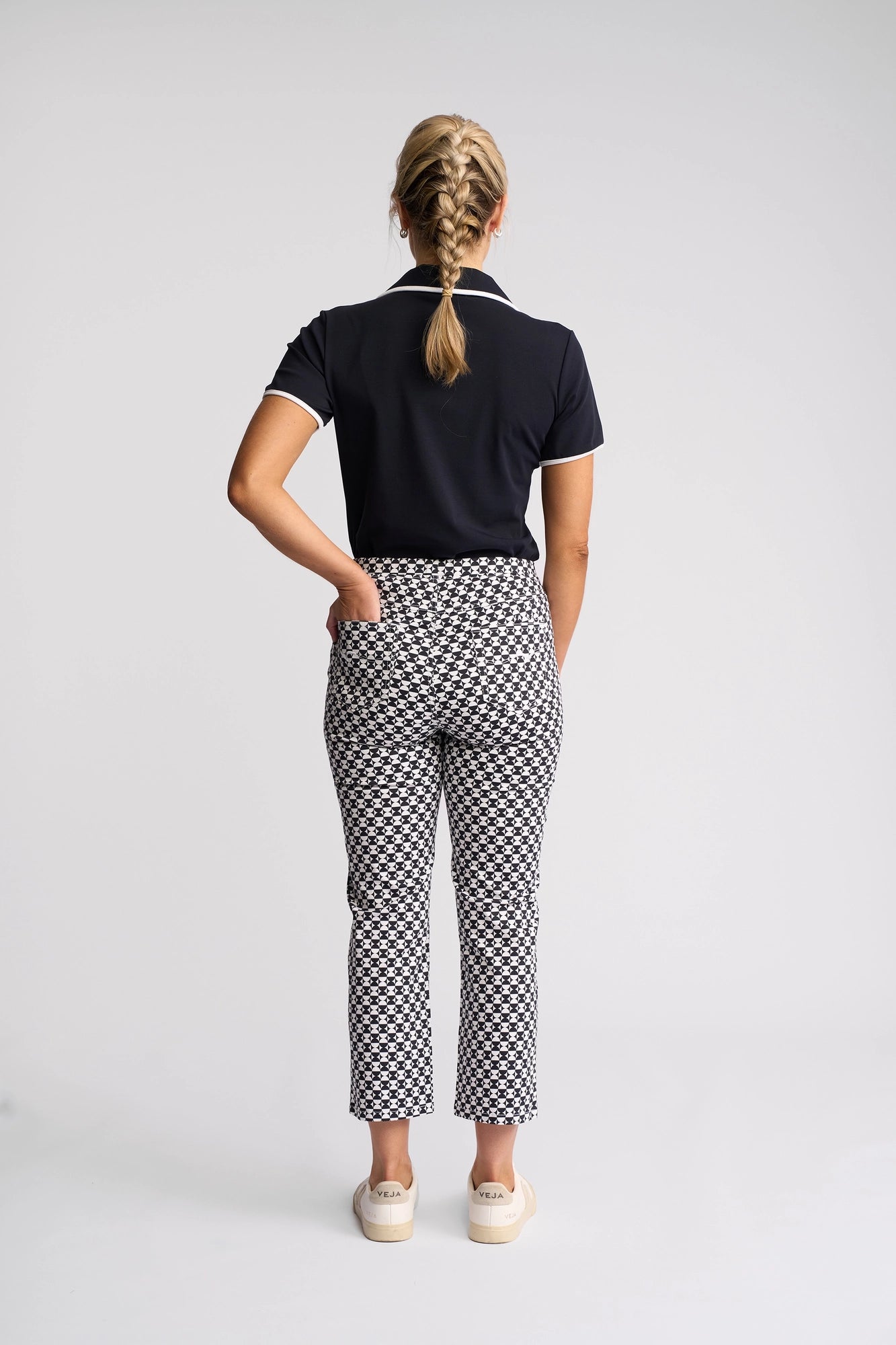A woman with blonde braided hair stands facing away from the camera, wearing a black collared shirt, Vassalli’s Sierra 7/8 Pant - Dixie slim-leg pants made from stretch cotton, and white shoes. She has one hand in her pocket against a plain white background.