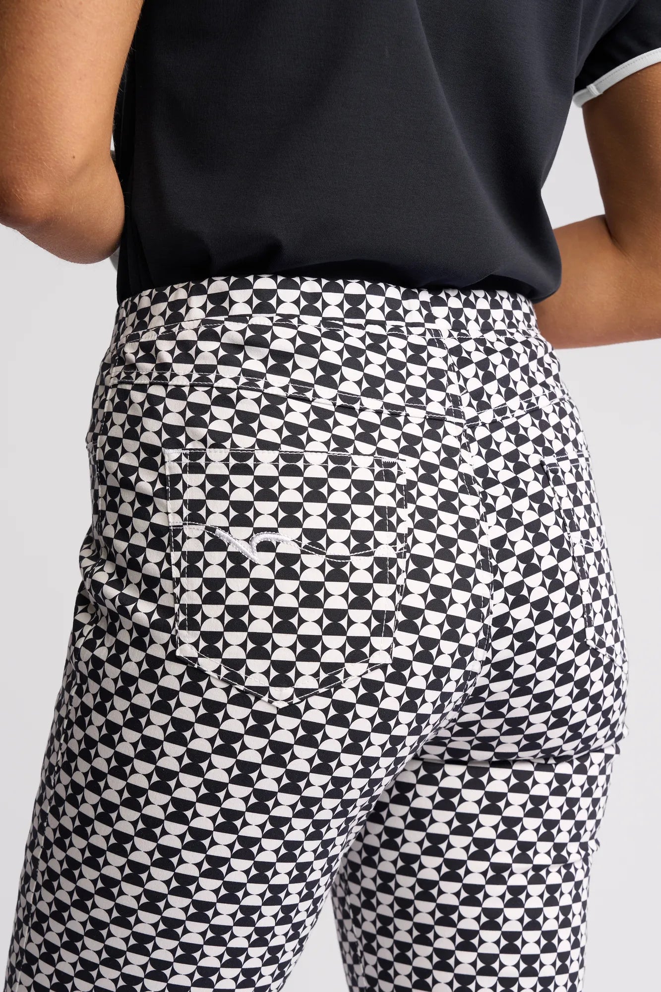 A person wearing the Vassalli Sierra 7/8 Pant - Dixie, featuring a black and white geometric pattern in stretch cotton with a slim-leg fit, styled with a black shirt, shown from the waist down and viewed from behind.