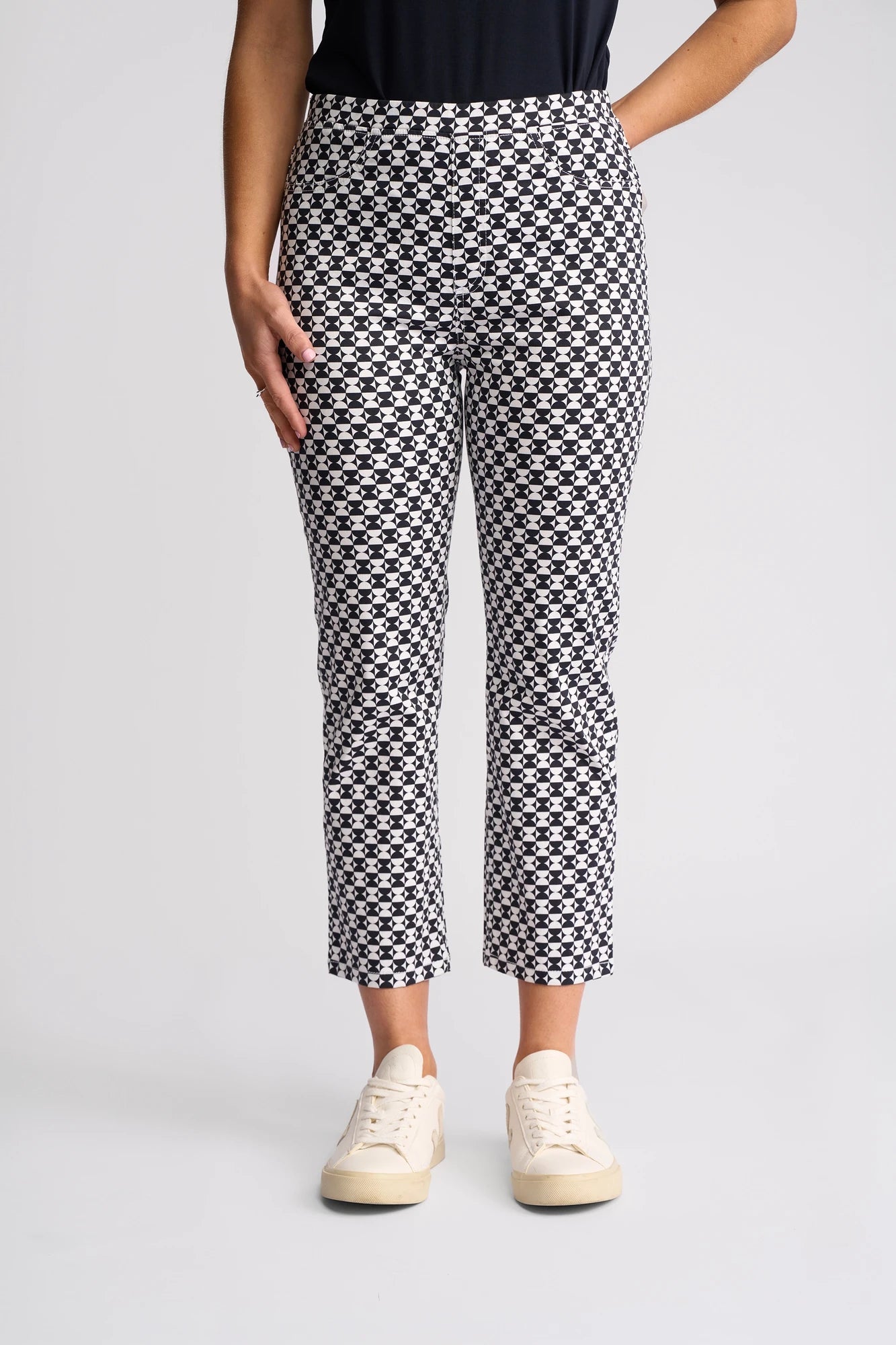 A person wearing Vassalli’s Sierra 7/8 Pant - Dixie, featuring a black-and-white checkered print in stretch cotton, paired with cream sneakers and a black top. The image is cropped from mid-torso to feet against a plain light gray background.