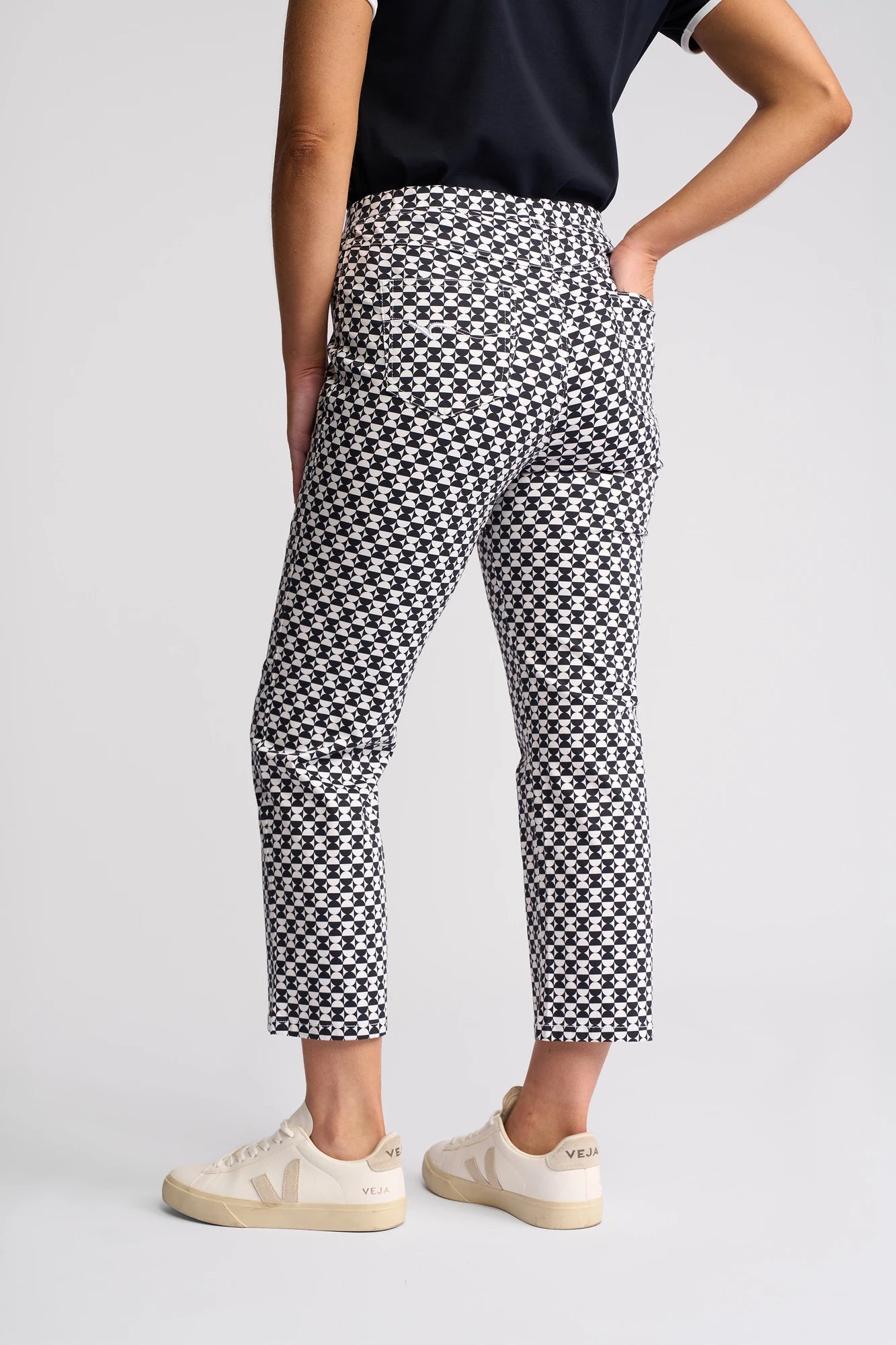 A person stands with one hand on their hip, wearing the Sierra 7/8 Pant - Dixie by Vassalli in a black and white checkered slim-leg style crafted from stretch cotton, paired with a black short-sleeve shirt and beige Veja sneakers against a plain light background.