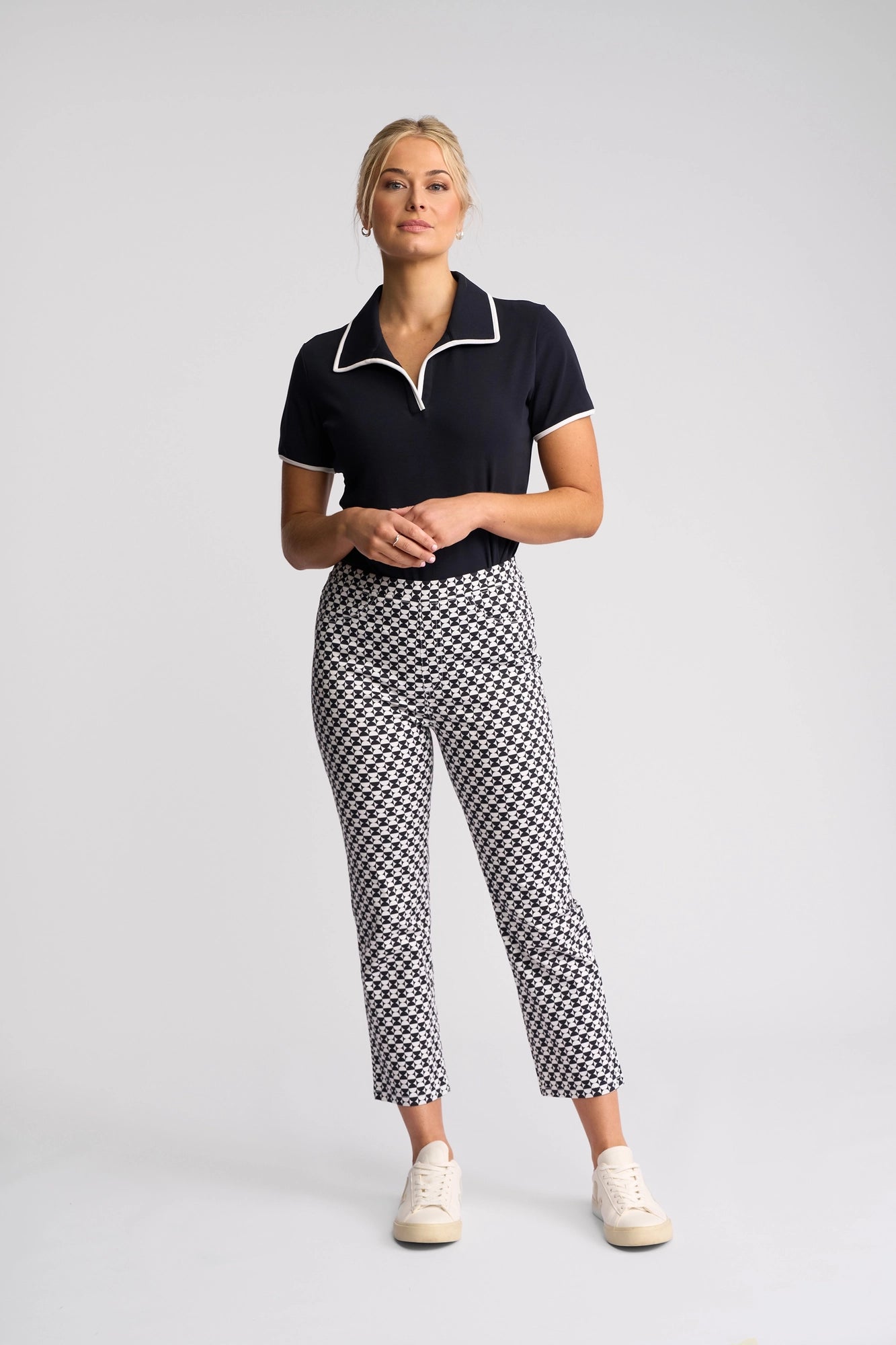 A woman stands against a light gray background wearing a black short-sleeve collared shirt, Vassalli’s Sierra 7/8 Pant - Dixie with an eye-catching pattern, and white sneakers. Her blonde hair is pulled back as she looks slightly upward, hands clasped.