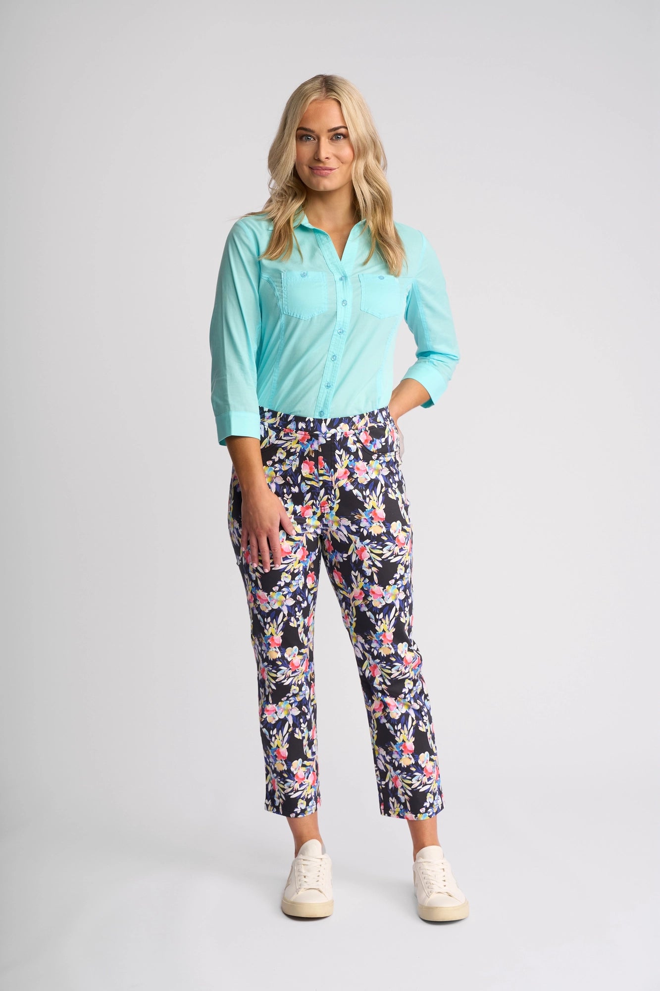 A woman with long blonde hair wears a light blue button-up shirt, Vassalli Sierra 7/8 Pant - Lani, and white sneakers, standing against a plain light gray background.