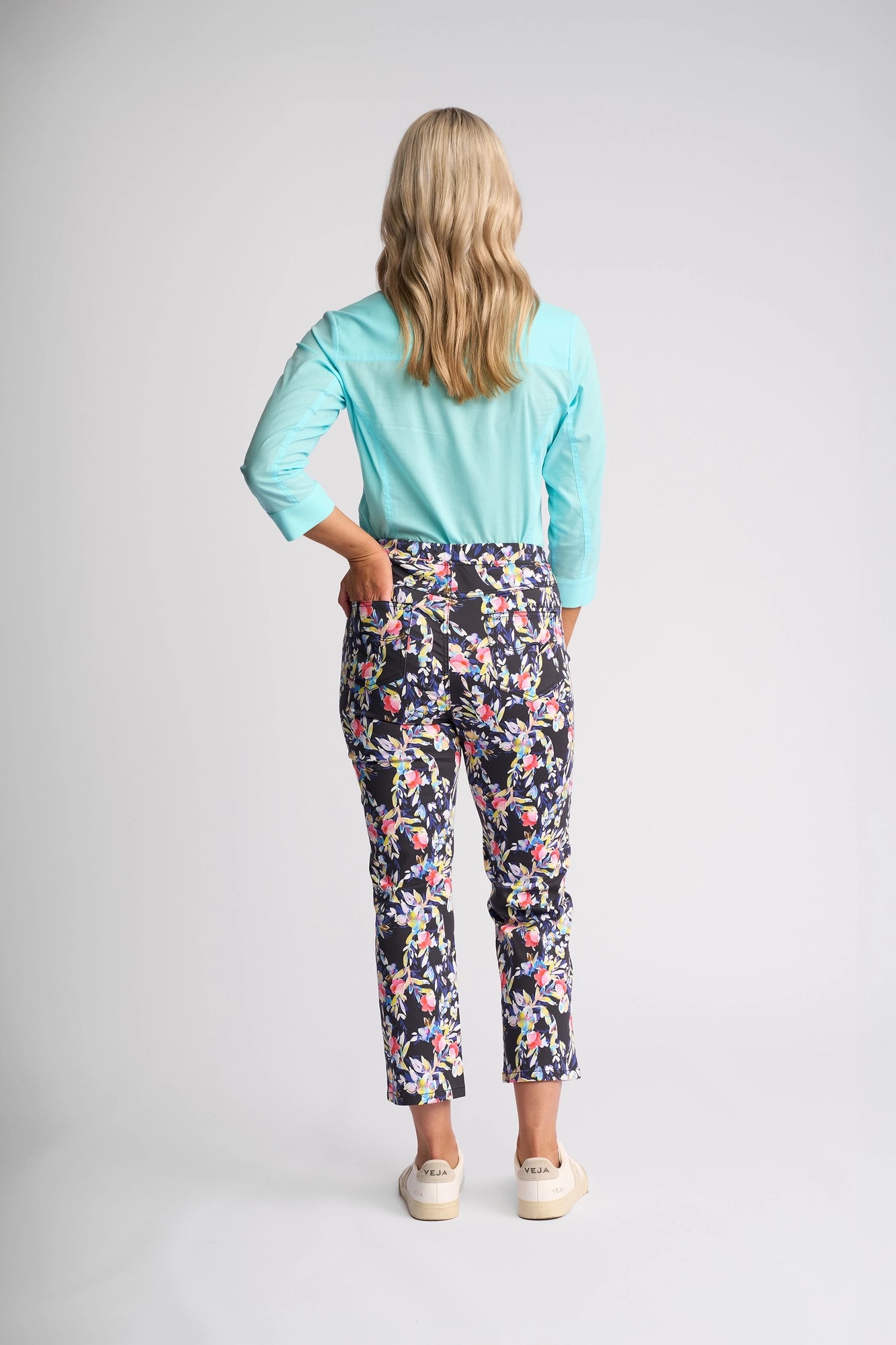 A person with long blonde hair stands facing away, wearing a light blue top and the Vassalli Sierra 7/8 Pant - Lani: floral patterned, slim-leg pull-on pants made from stretch cotton, paired with white shoes against a plain light background.