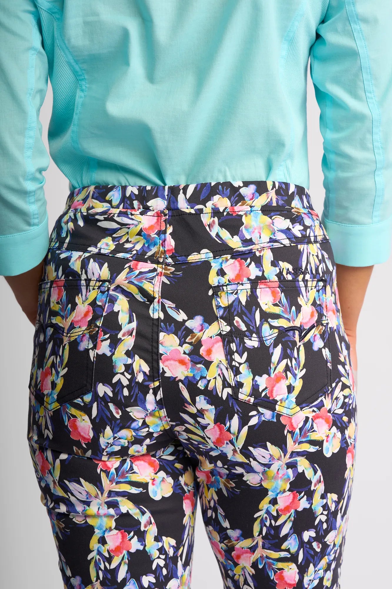 A person wearing a light blue shirt and Vassalli’s Sierra 7/8 Pant - Lani is shown from the back, with the focus on the waist and upper legs. These pull-on pants feature a colorful floral pattern on a dark background.