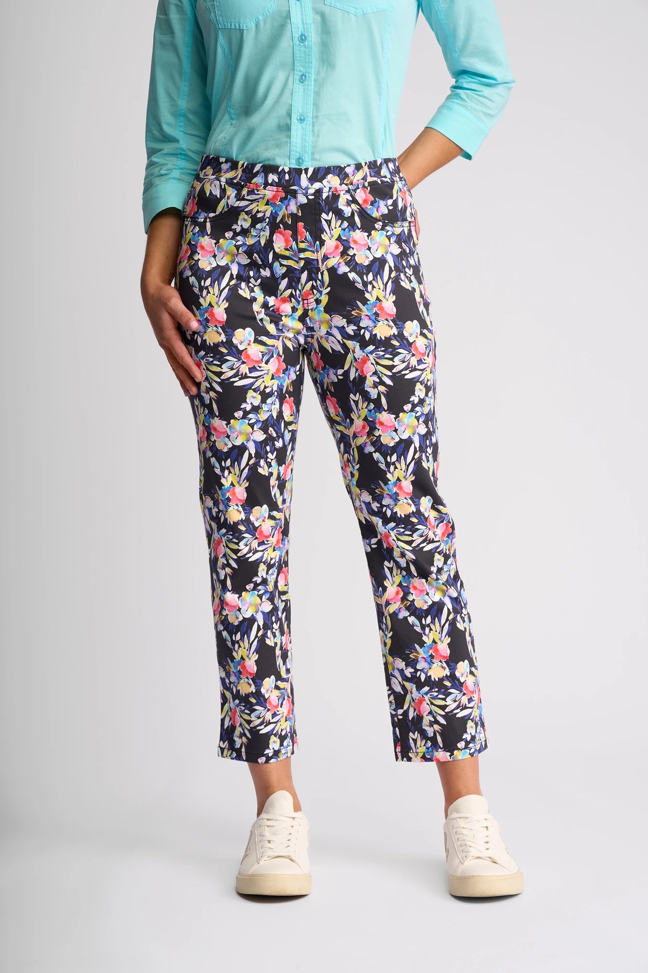 A person is wearing a light blue long-sleeve shirt and Vassalli’s Sierra 7/8 Pant - Lani, featuring a colorful floral pattern in stretch cotton, paired with white sneakers. Only the body is visible; the head is not shown.