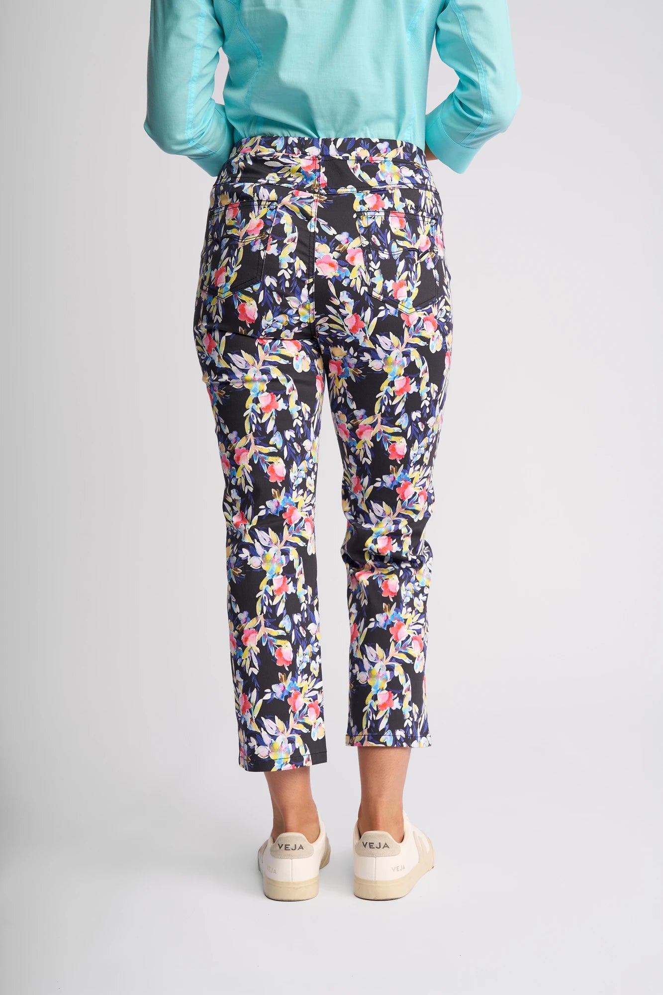 A person stands with their back to the camera, wearing a turquoise shirt and Vassalli Sierra 7/8 Pant - Lani, a pull-on style with slim legs crafted from stretch cotton and adorned with pink, white, and purple floral prints. They finish the look with cream-colored sneakers featuring VEJA on the heels.