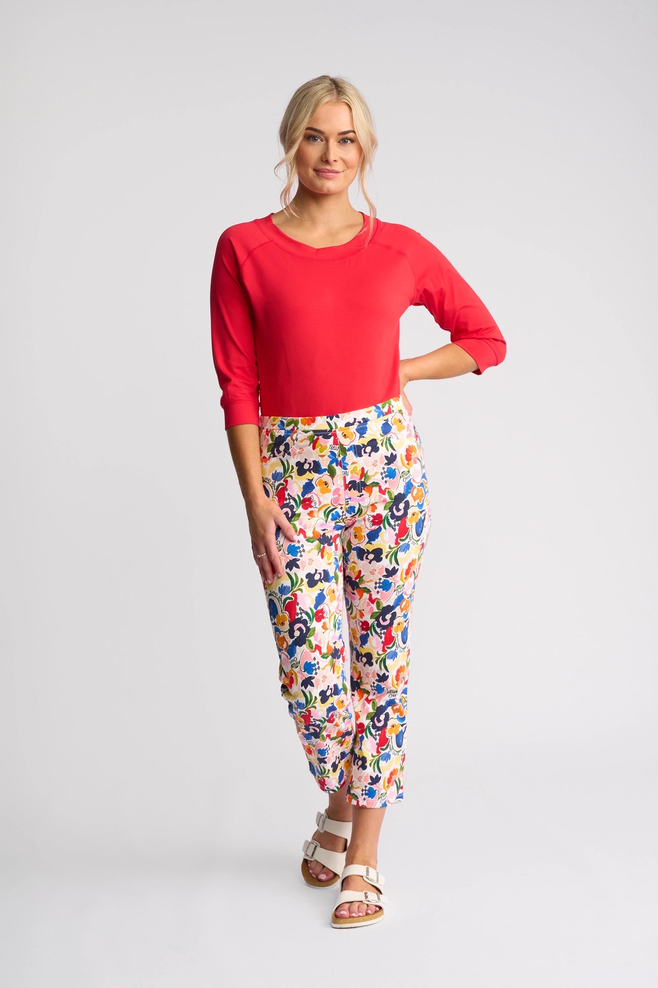 Woman standing against a plain background, smiling with one hand on her hip, wearing a bright red top, Vassalli Sierra 7/8 Pant - Mon Cheri in a vibrant floral print stretch cotton, and white sandals.
