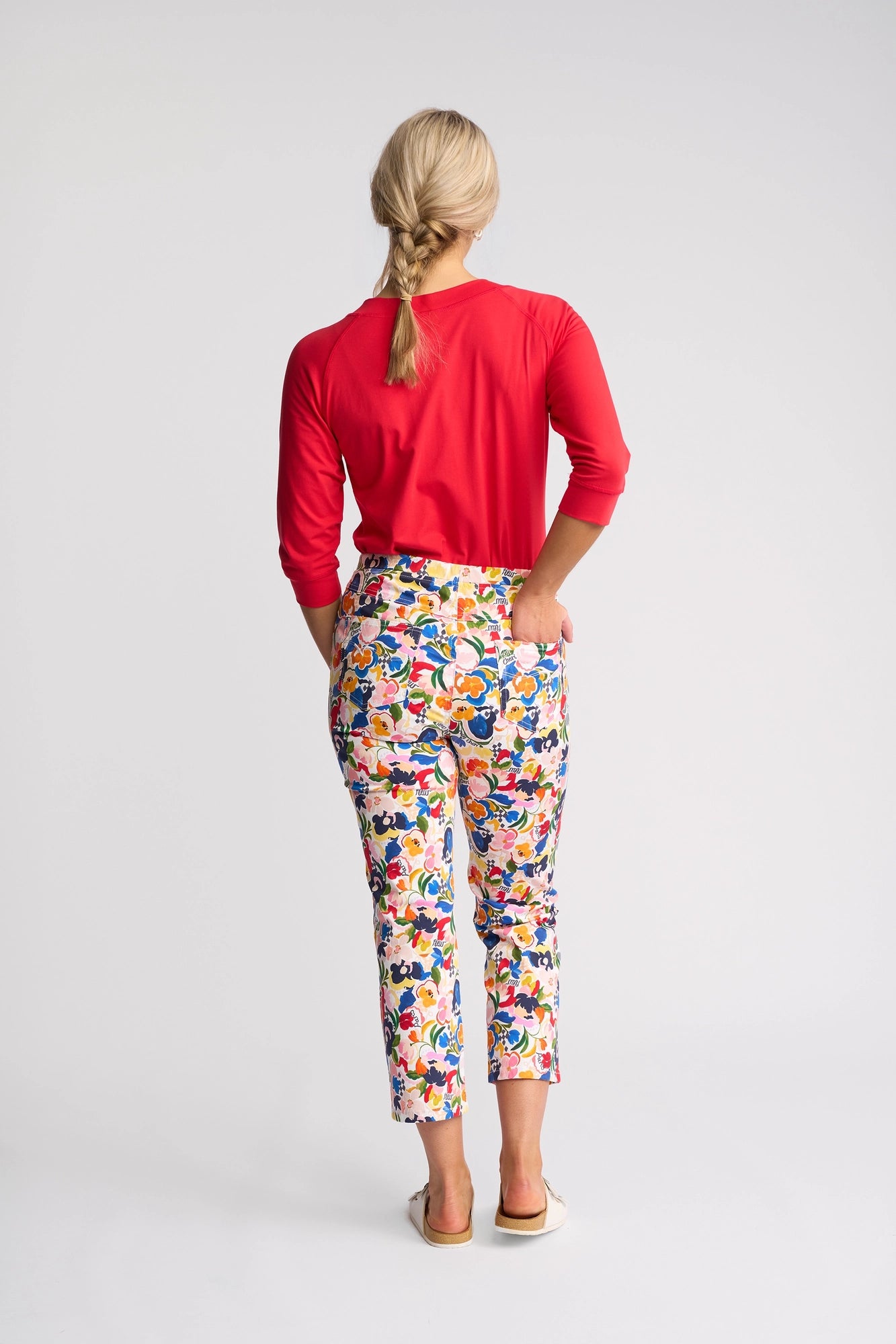 A woman stands facing away, wearing a red long-sleeve shirt and the Vassalli Sierra 7/8 Pant - Mon Cheri, featuring a colorful floral print. Her blonde hair is in a braid and she wears flat shoes, set against a plain white background.