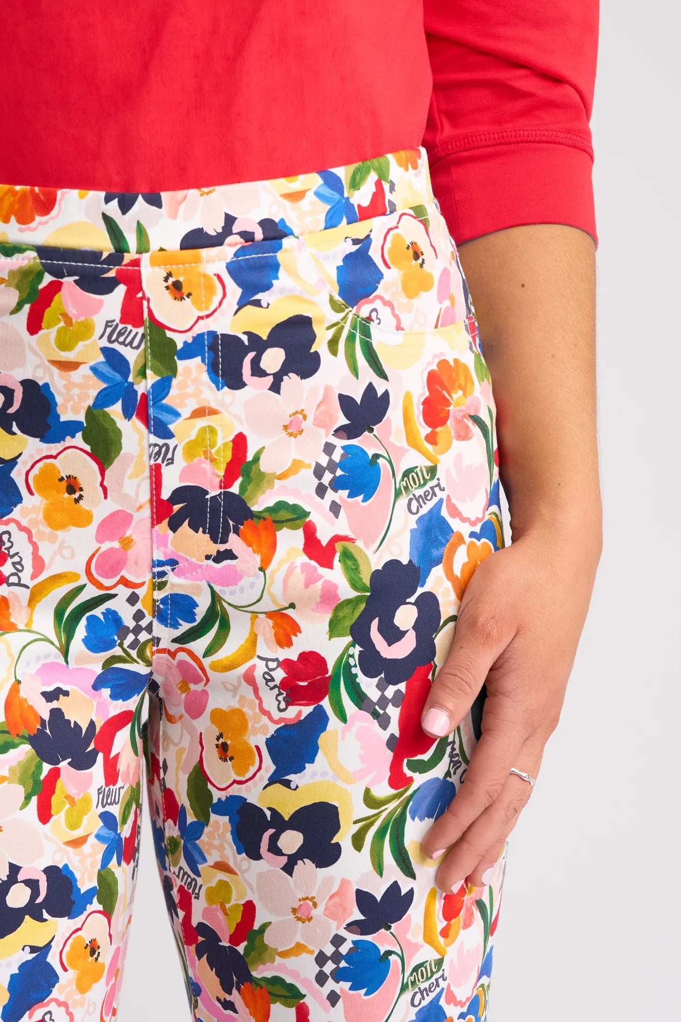 A person wearing a red top and vibrant Vassalli Sierra 7/8 Pant - Mon Cheri, featuring a floral print with various flowers and “oui cherie” text. Their right hand rests by their side, displaying a delicate ring on one finger.