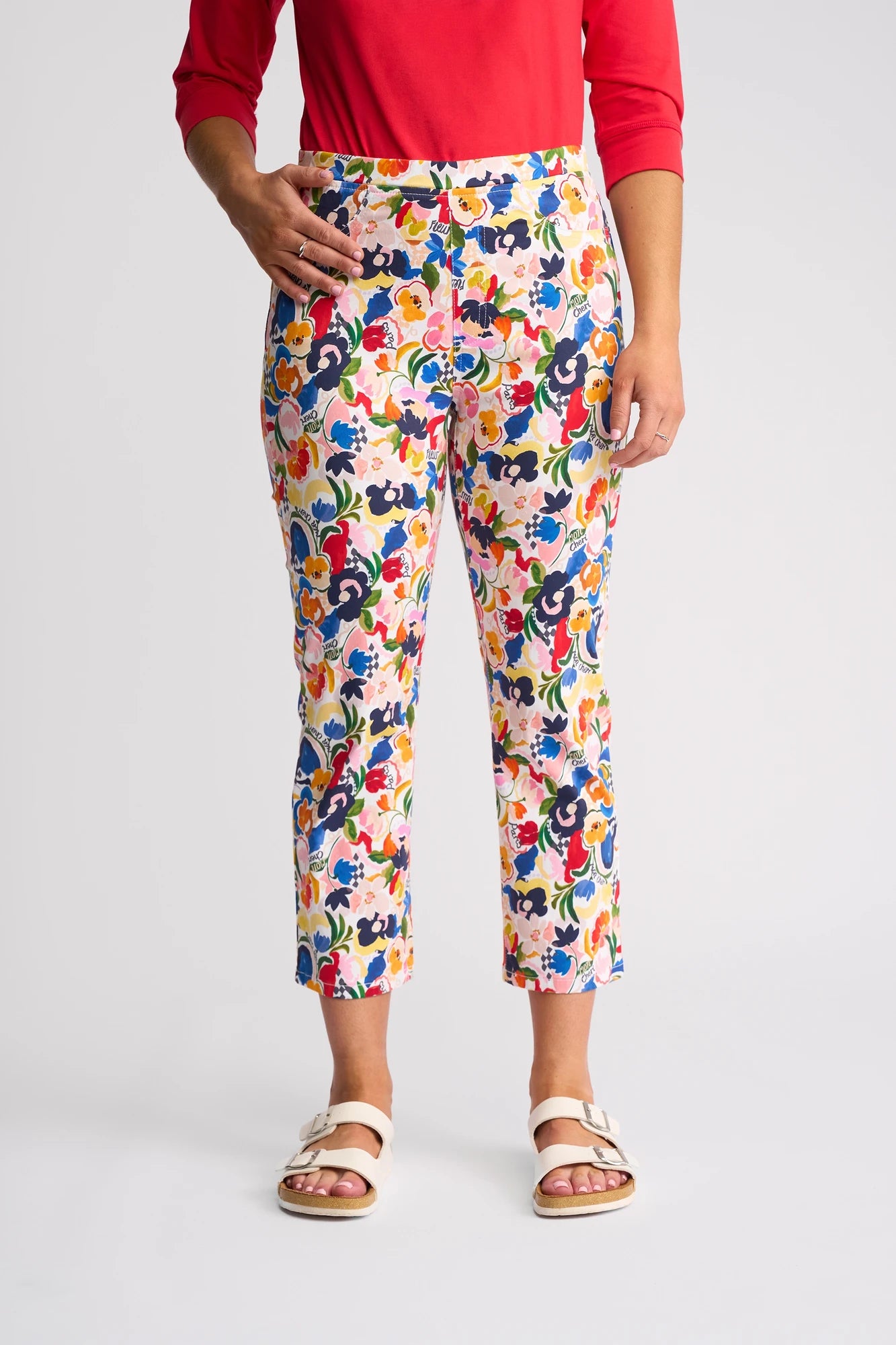 A person wearing a red top, the Vassalli Sierra 7/8 Pant - Mon Cheri (a colorful stretch cotton floral print pant), and white sandals stands on a plain, light background. Their left hand rests on their hip.