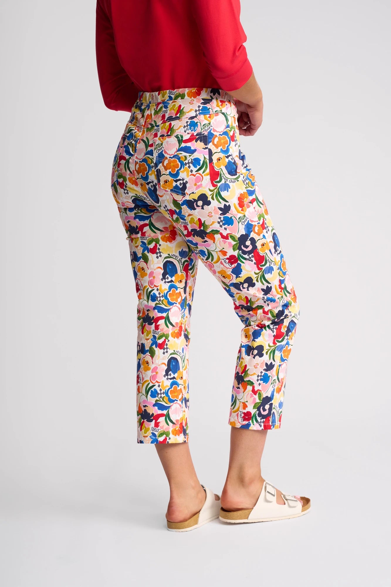 A person wearing vibrant Vassalli Sierra 7/8 Pant - Mon Cheri, paired with a red top and light sandals, stands against a plain background, facing away at an angle.
