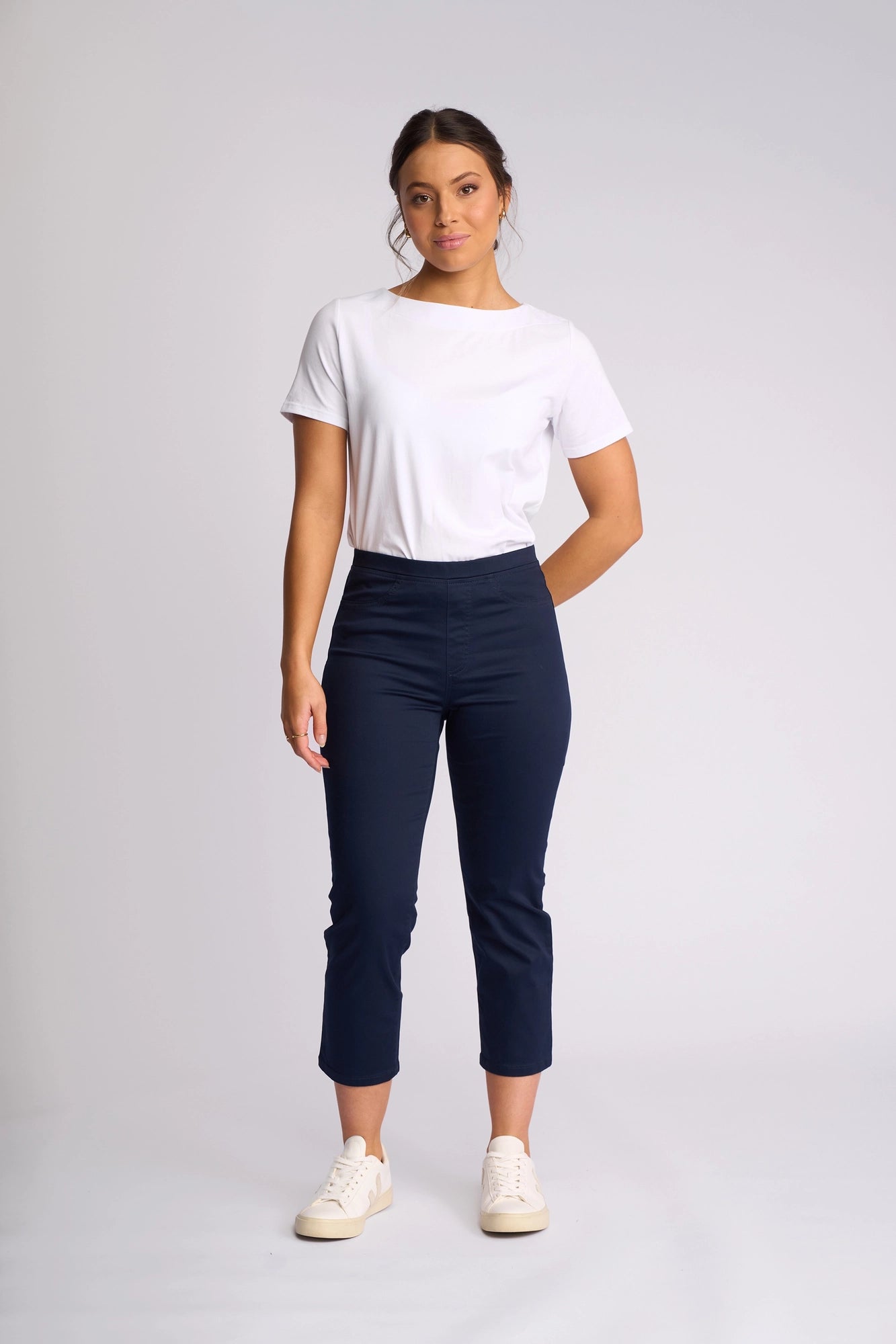 A woman stands against a plain light background, wearing a white t-shirt and the Vassalli Sierra 7/8 Pant in Navy—stylish, slim-leg, ankle-length pull-on pants crafted from stretch cotton for all-day comfort. She pairs them with white sneakers and poses with one hand behind her back.