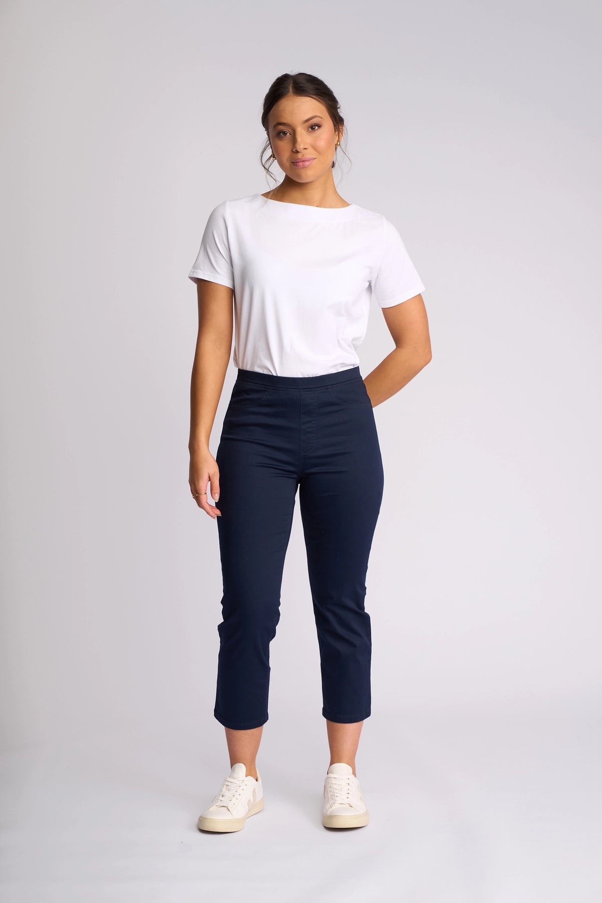 A woman stands against a plain light background, wearing a white t-shirt and the Vassalli Sierra 7/8 Pant in Navy—stylish, slim-leg, ankle-length pull-on pants crafted from stretch cotton for all-day comfort. She pairs them with white sneakers and poses with one hand behind her back.