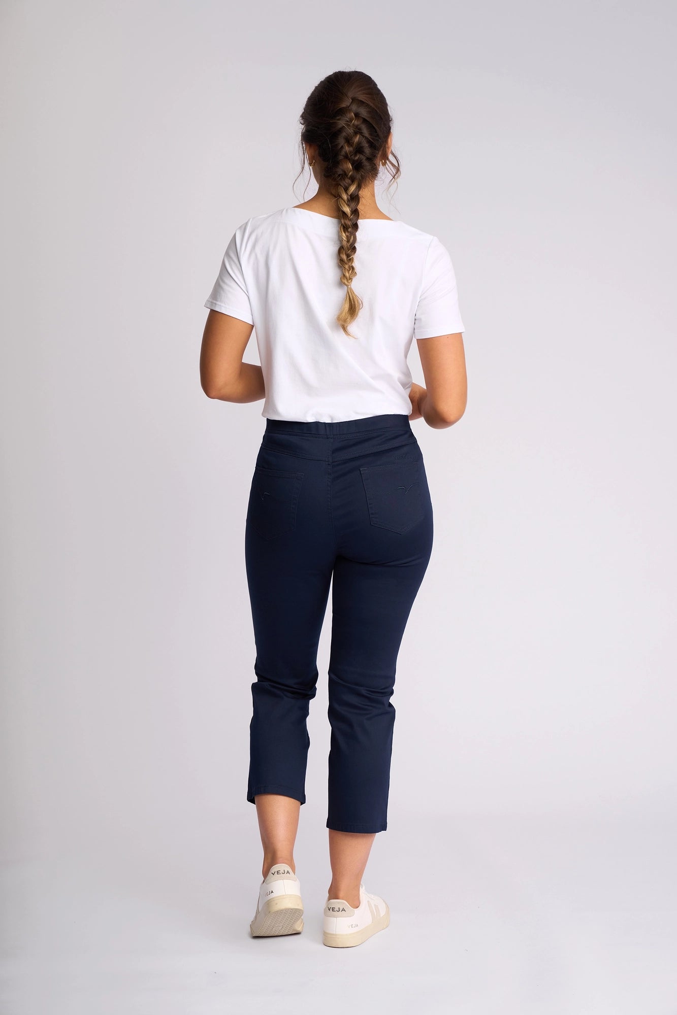 A woman with braided hair stands facing away against a plain light background, wearing a white t-shirt, white sneakers, and Vassalli’s Sierra 7/8 Pant in Navy.