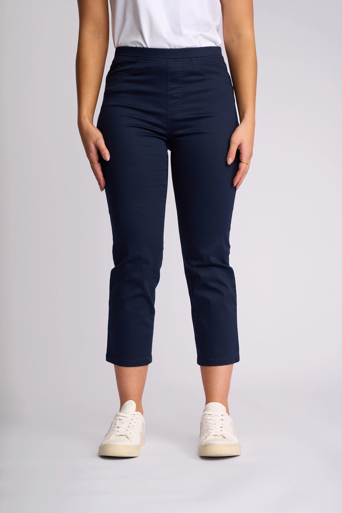 A person wearing a white t-shirt, Vassalli Sierra 7/8 Pant in navy crafted from stretch cotton, and white sneakers stands facing forward against a plain light background.