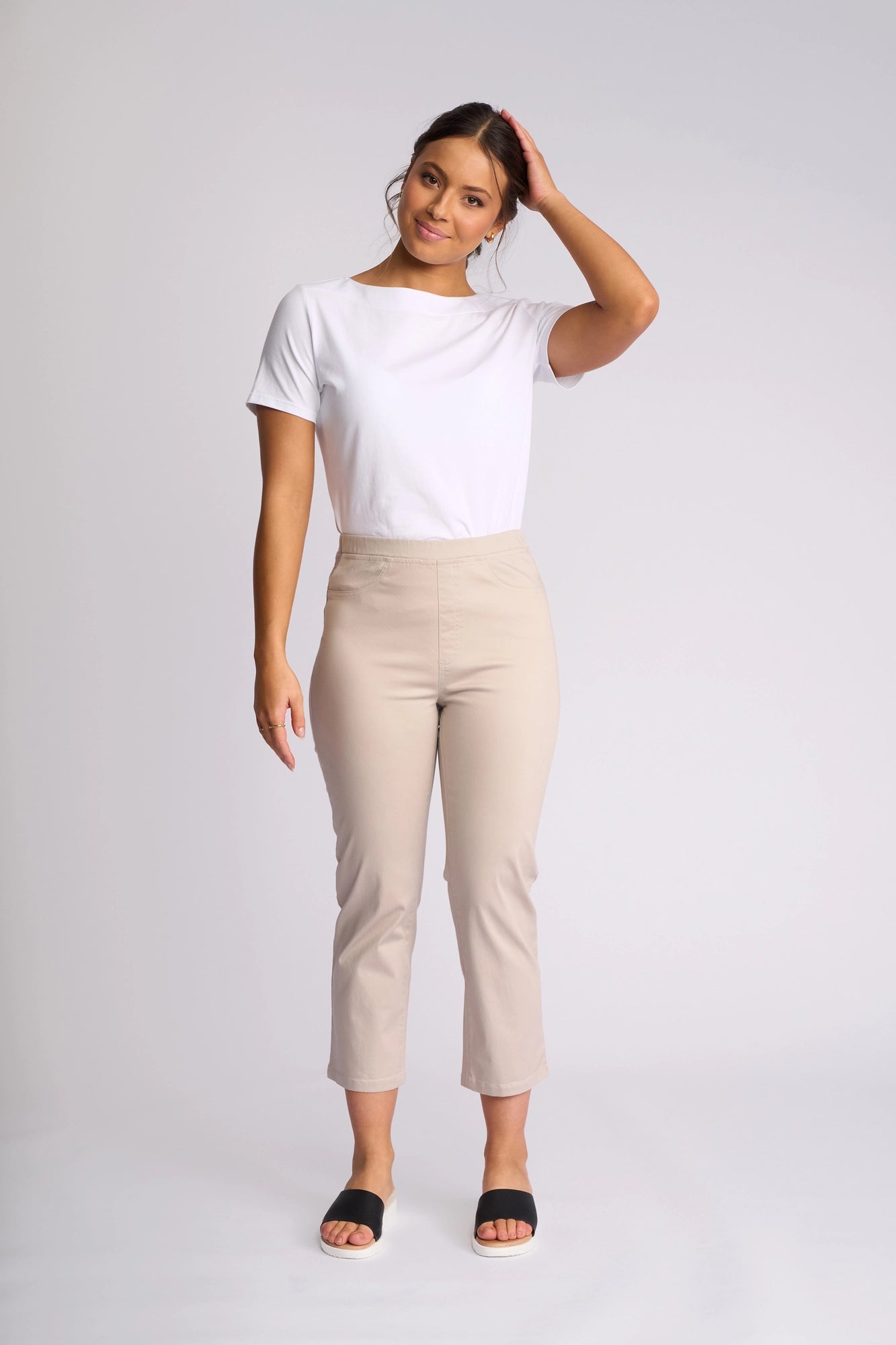 A woman stands against a plain background, wearing a white t-shirt, Vassalli Sierra 7/8 Pant in Pumice with a slim-leg fit crafted from stretch cotton, and black sandals. She has one hand touching her hair and is smiling gently.