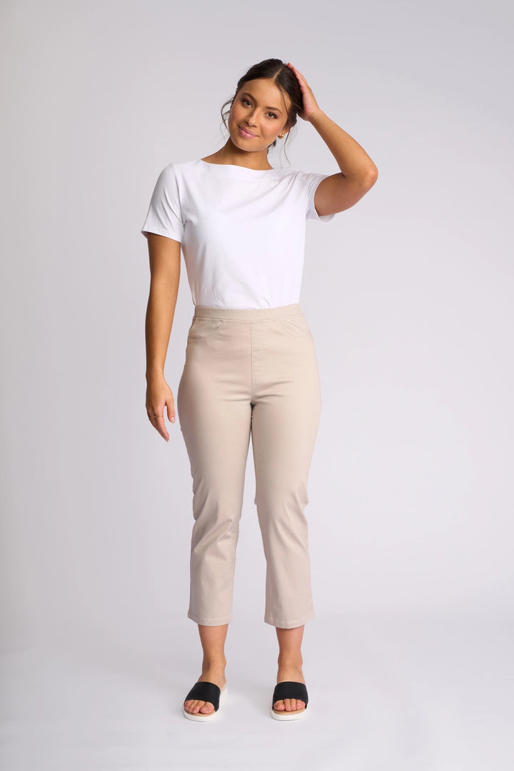 A woman stands against a plain background, wearing a white t-shirt, Vassalli Sierra 7/8 Pant in Pumice with a slim-leg fit crafted from stretch cotton, and black sandals. She has one hand touching her hair and is smiling gently.
