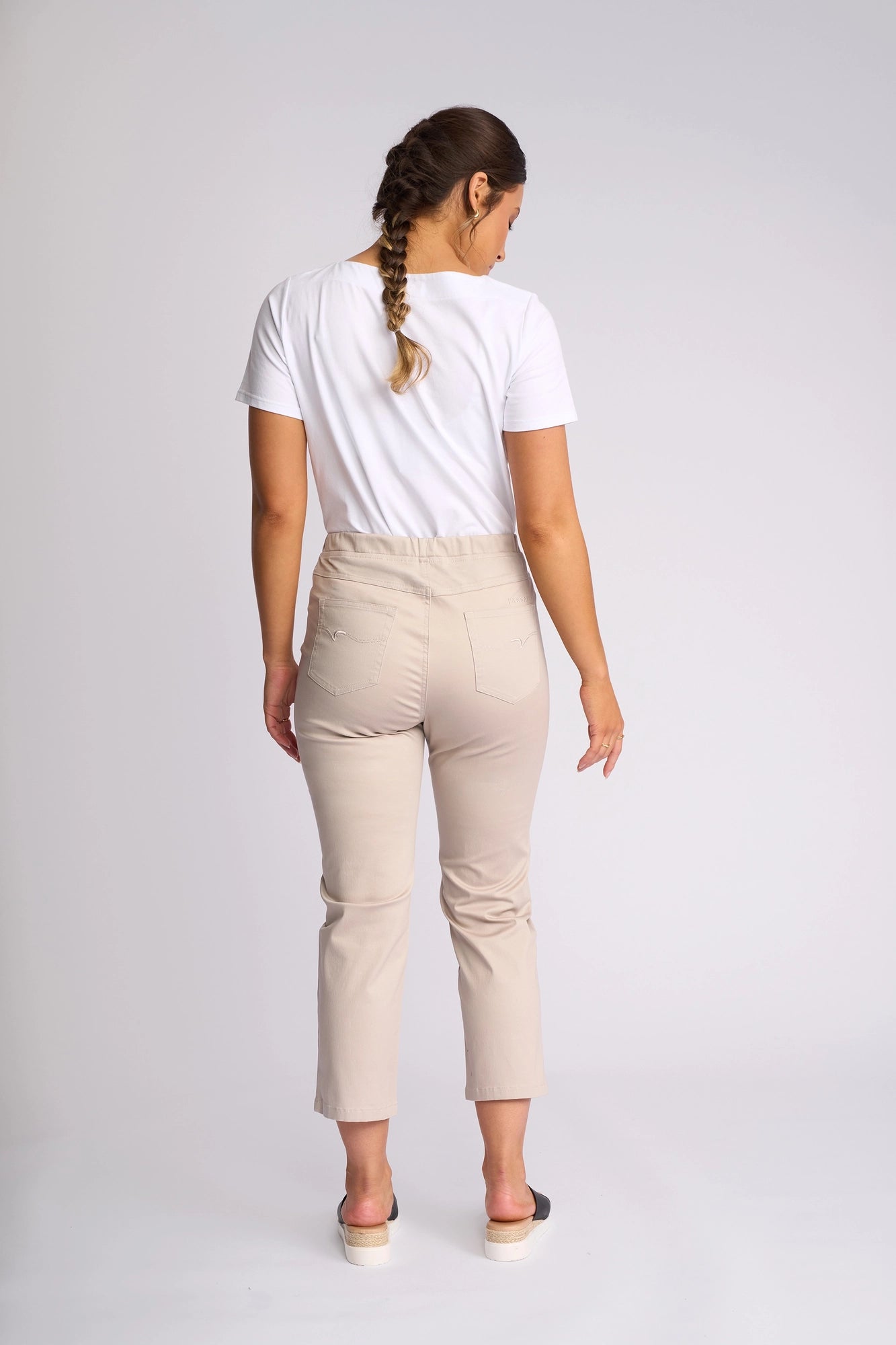 A woman with a braid stands and faces away in a studio with a plain light background, wearing a white t-shirt, the Vassalli Sierra 7/8 Pant in Pumice, and black slip-on shoes.