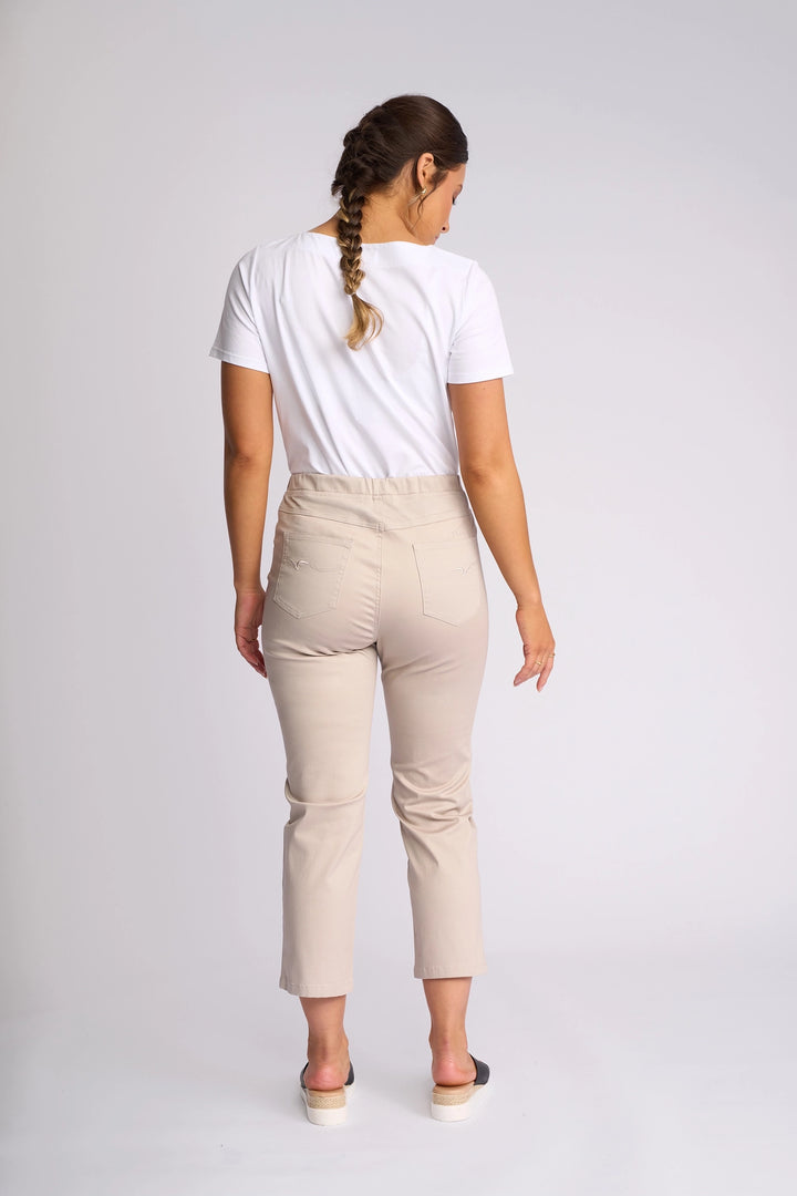 A woman with a braid stands and faces away in a studio with a plain light background, wearing a white t-shirt, the Vassalli Sierra 7/8 Pant in Pumice, and black slip-on shoes.