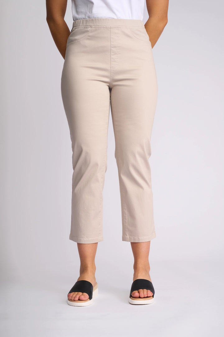 A person wearing the Vassalli Sierra 7/8 Pant in Pumice—beige slim-leg cropped pants crafted from stretch cotton—with a white shirt and black slide sandals, stands with hands behind their back against a plain light background.
