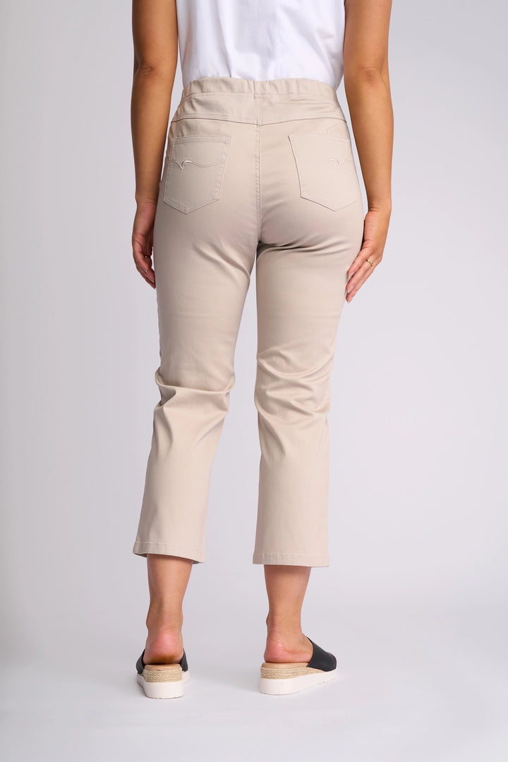 A person stands facing away against a plain light background, wearing Vassalli’s Sierra 7/8 Pant in Pumice—beige stretch cotton slim-leg pants—paired with a white shirt and black slip-on shoes with espadrille soles.
