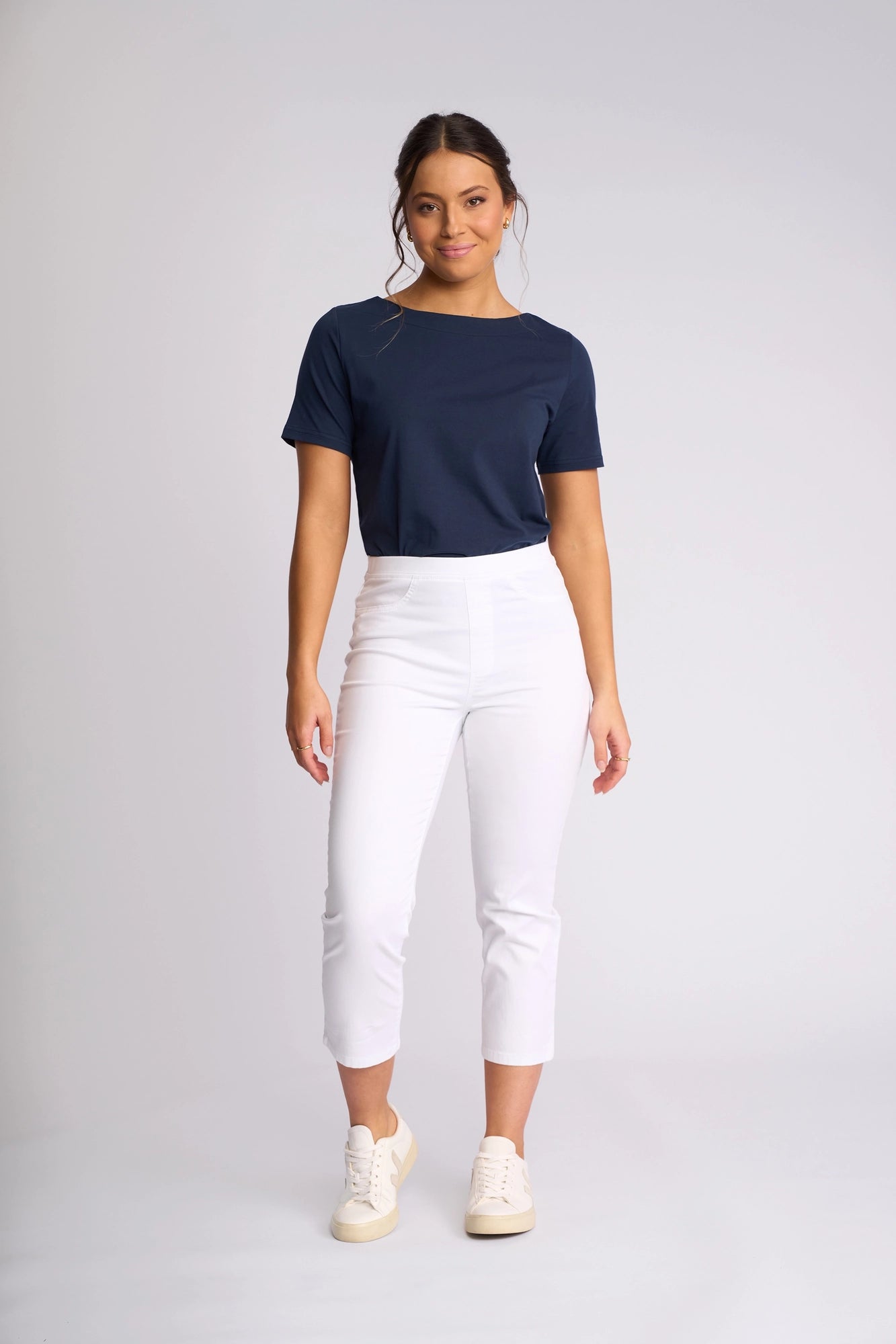 A woman stands facing forward, wearing a navy blue t-shirt and the Vassalli Sierra 7/8 Pant in White—crisp, slim-leg pull-on pants crafted from stretch cotton—paired with white sneakers, against a plain light background.