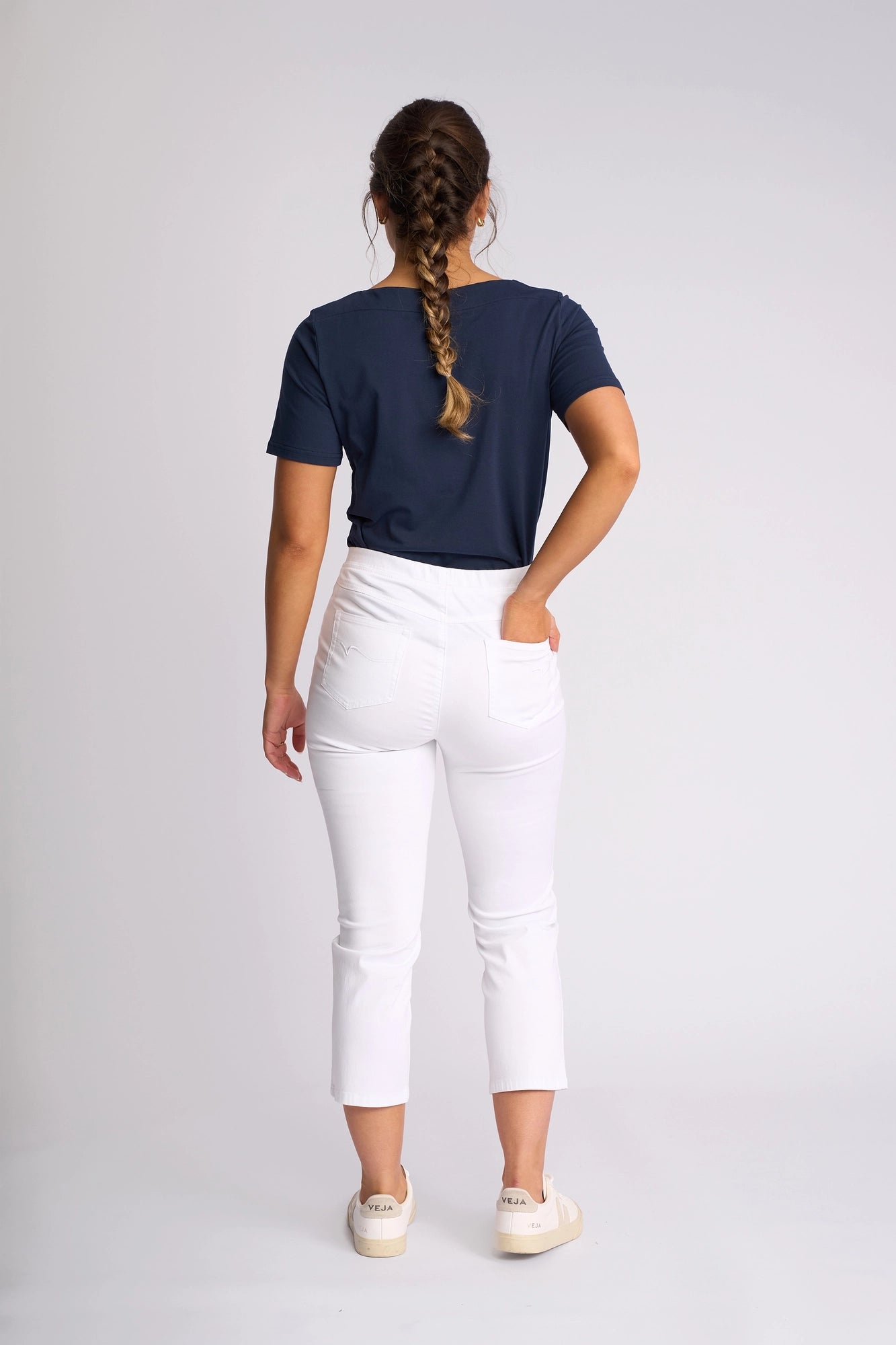 A woman with a braid stands facing away, wearing a navy T-shirt, white Vassalli Sierra 7/8 Pant, and white sneakers, with one hand in her pocket against a plain light background.
