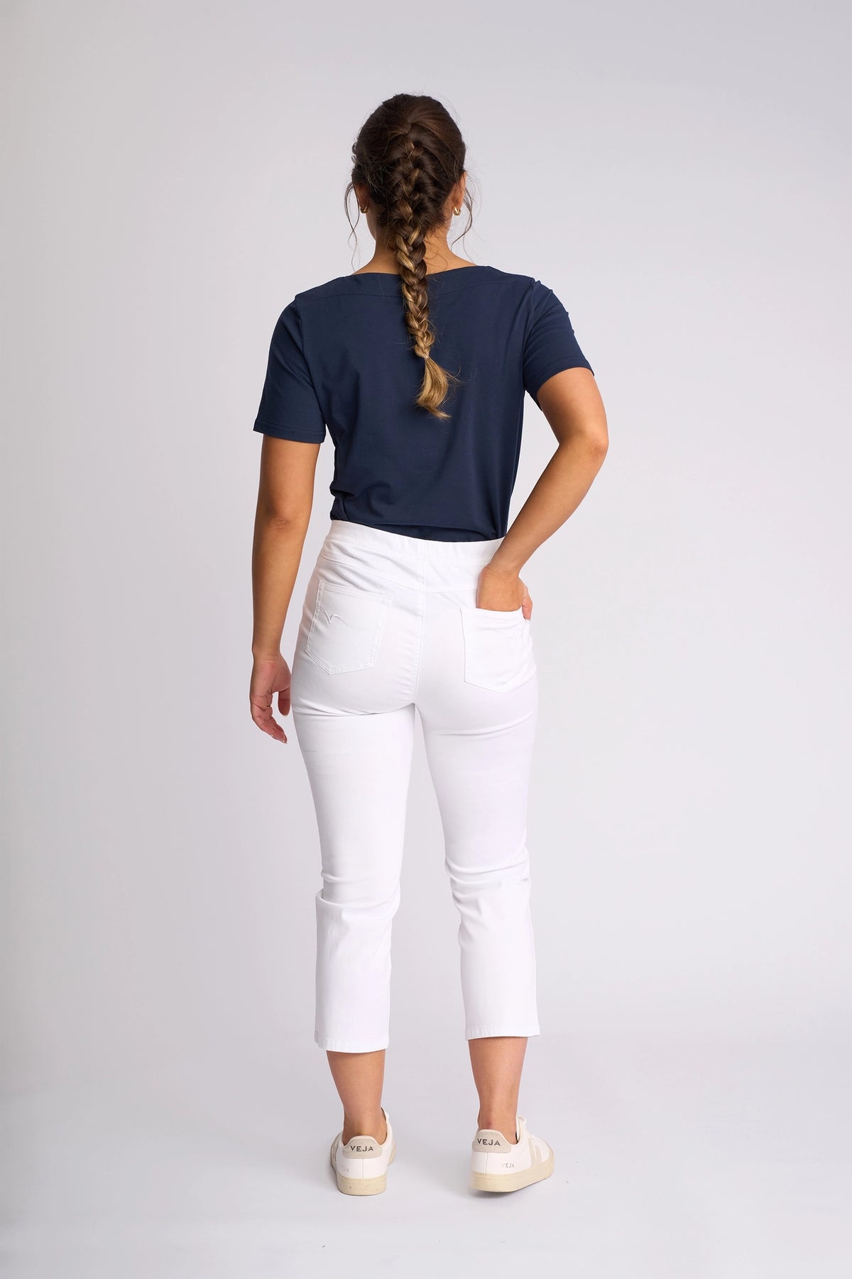 A woman with a braid stands facing away, wearing a navy T-shirt, white Vassalli Sierra 7/8 Pant, and white sneakers, with one hand in her pocket against a plain light background.