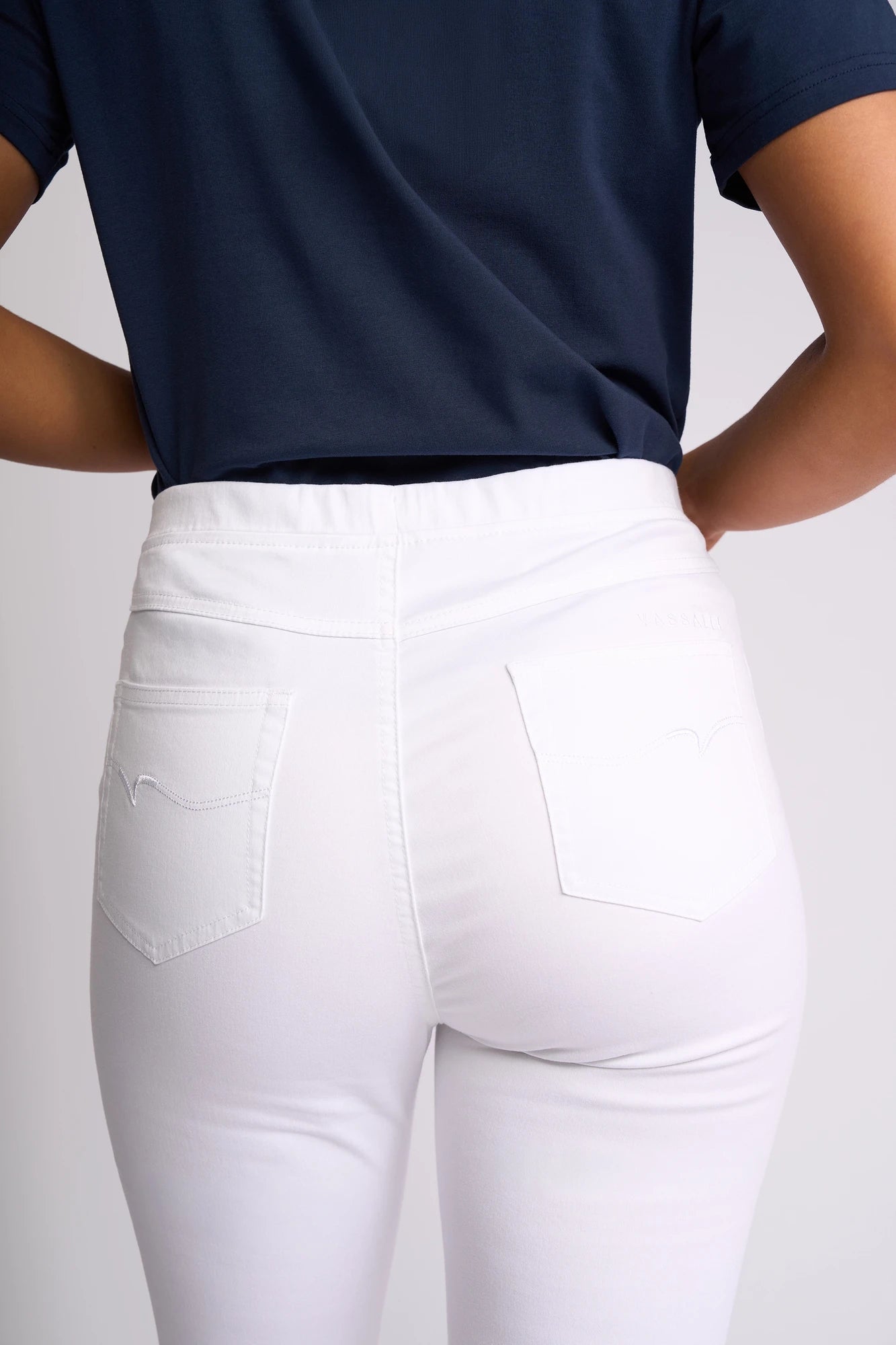 A person is shown from the back with their hands on their hips, wearing a navy blue shirt and the Vassalli Sierra 7/8 Pant in White. The image highlights the fit, stretch cotton fabric, and pockets of these slim-leg pull-on pants.