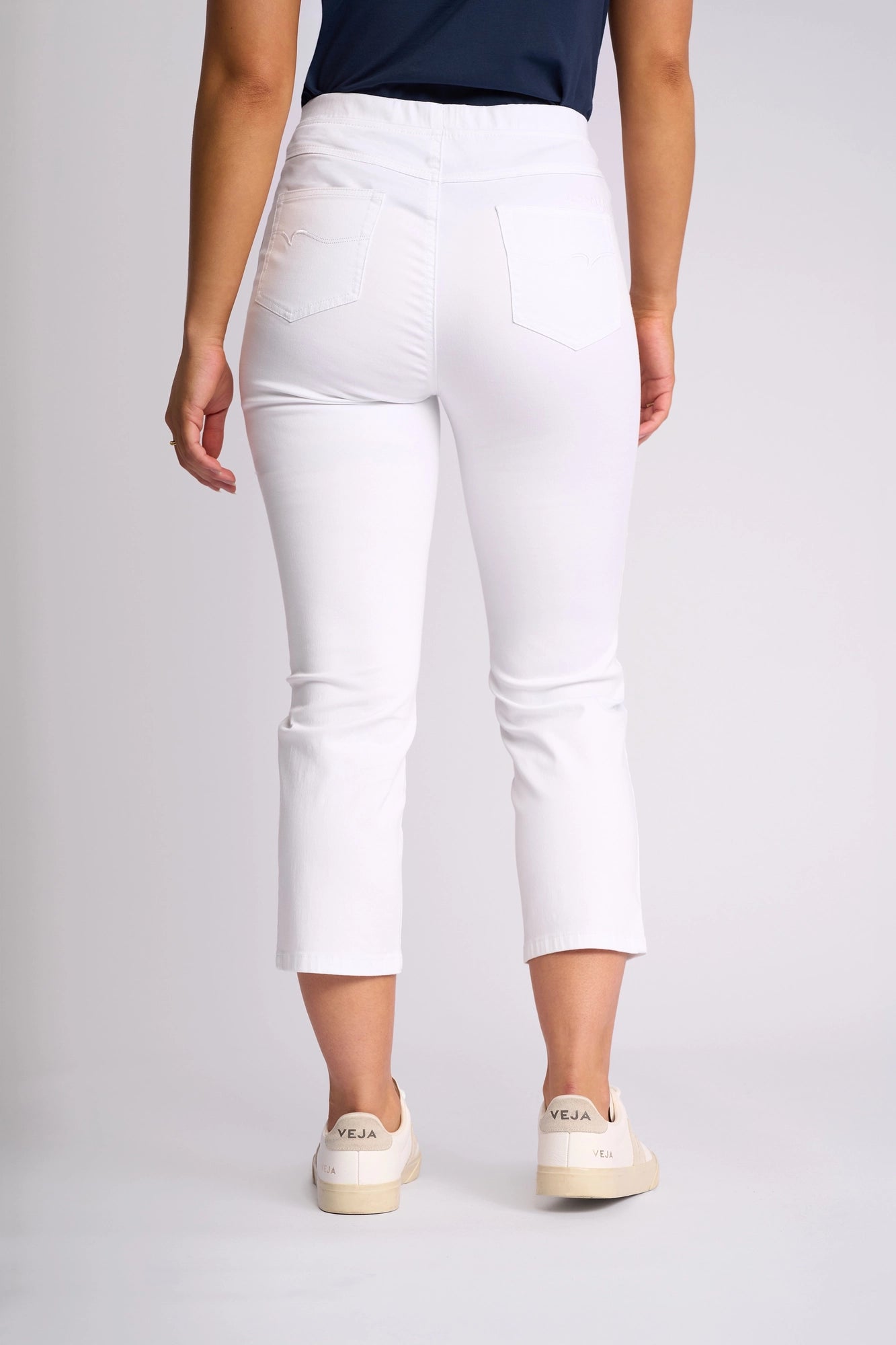 A person stands facing away, wearing the Vassalli Sierra 7/8 Pant in white—crafted from stretch cotton with a slim-leg pull-on design—paired with a navy top and beige Veja sneakers, photographed against a plain light background.