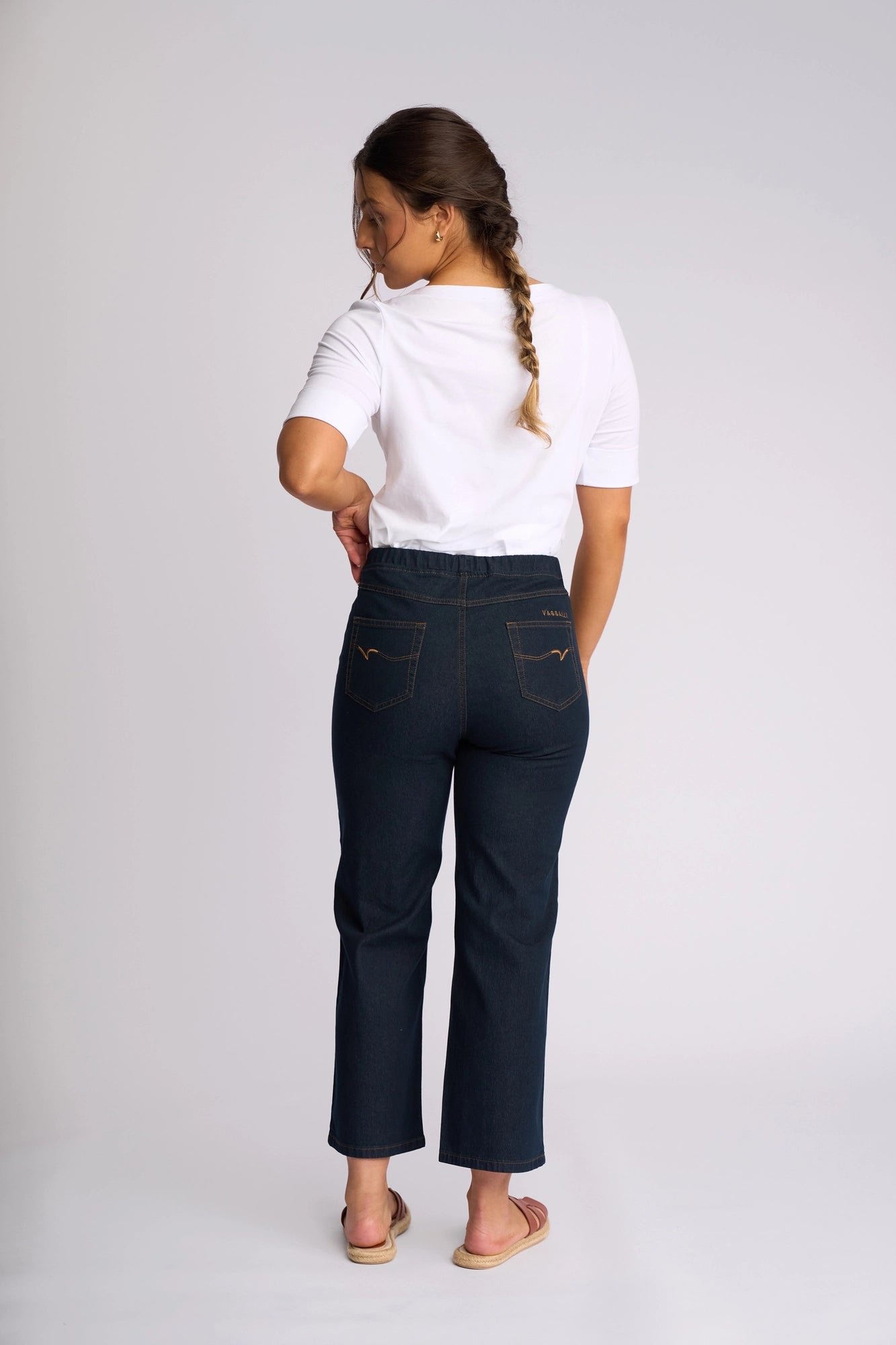 A woman stands facing away, wearing a white short-sleeve shirt, sandals, and the Vassalli Seattle Pull On - Indigo Denim jeans. Her hair is styled in a braid, and she stands against a plain light background.