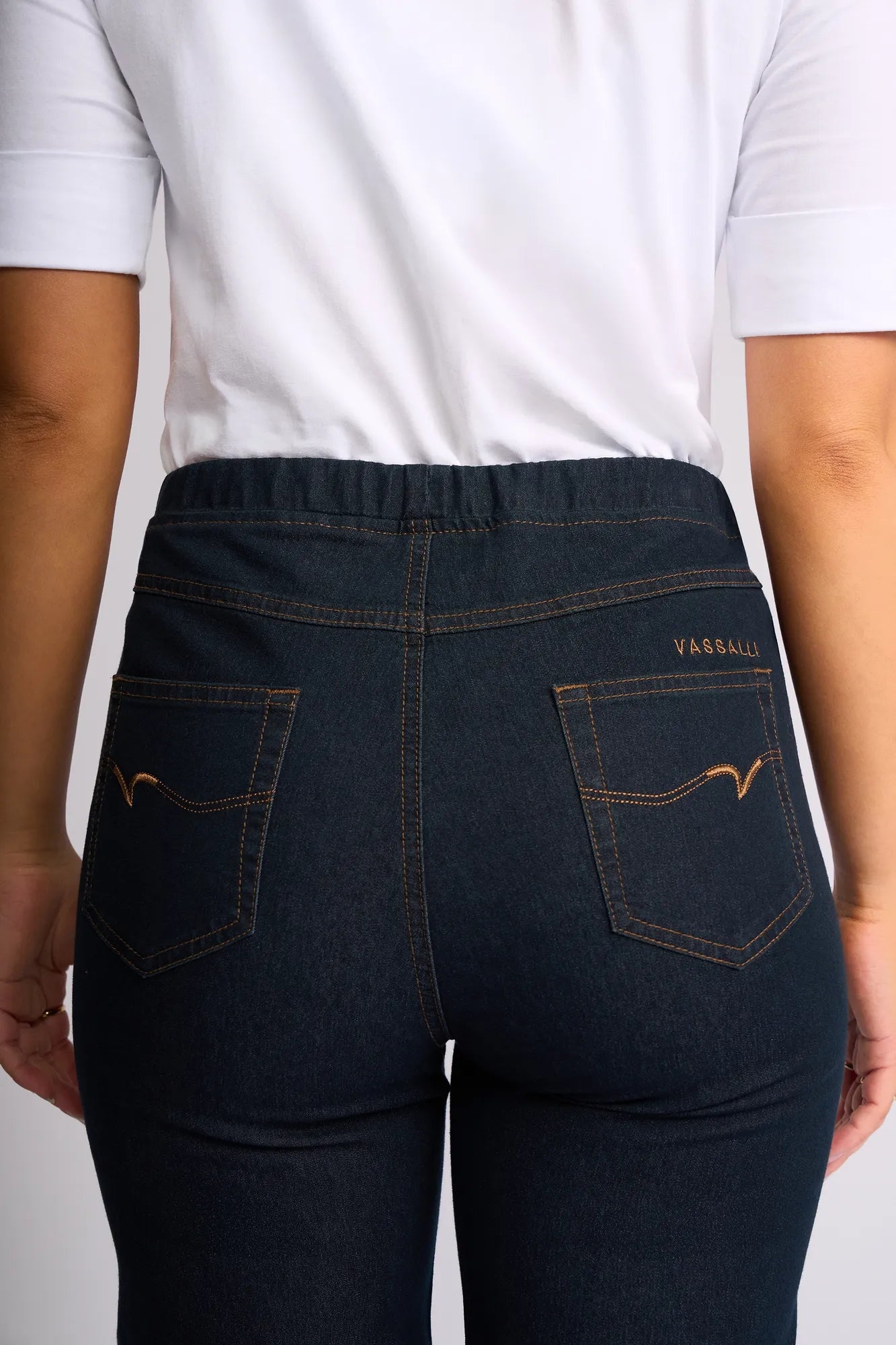 A person wearing a white shirt and Vassalli Seattle Pull On indigo denim jeans is seen from the back; the straight-leg jeans feature two back pockets with yellow stitching and “Vassalli” embroidered above the right pocket.