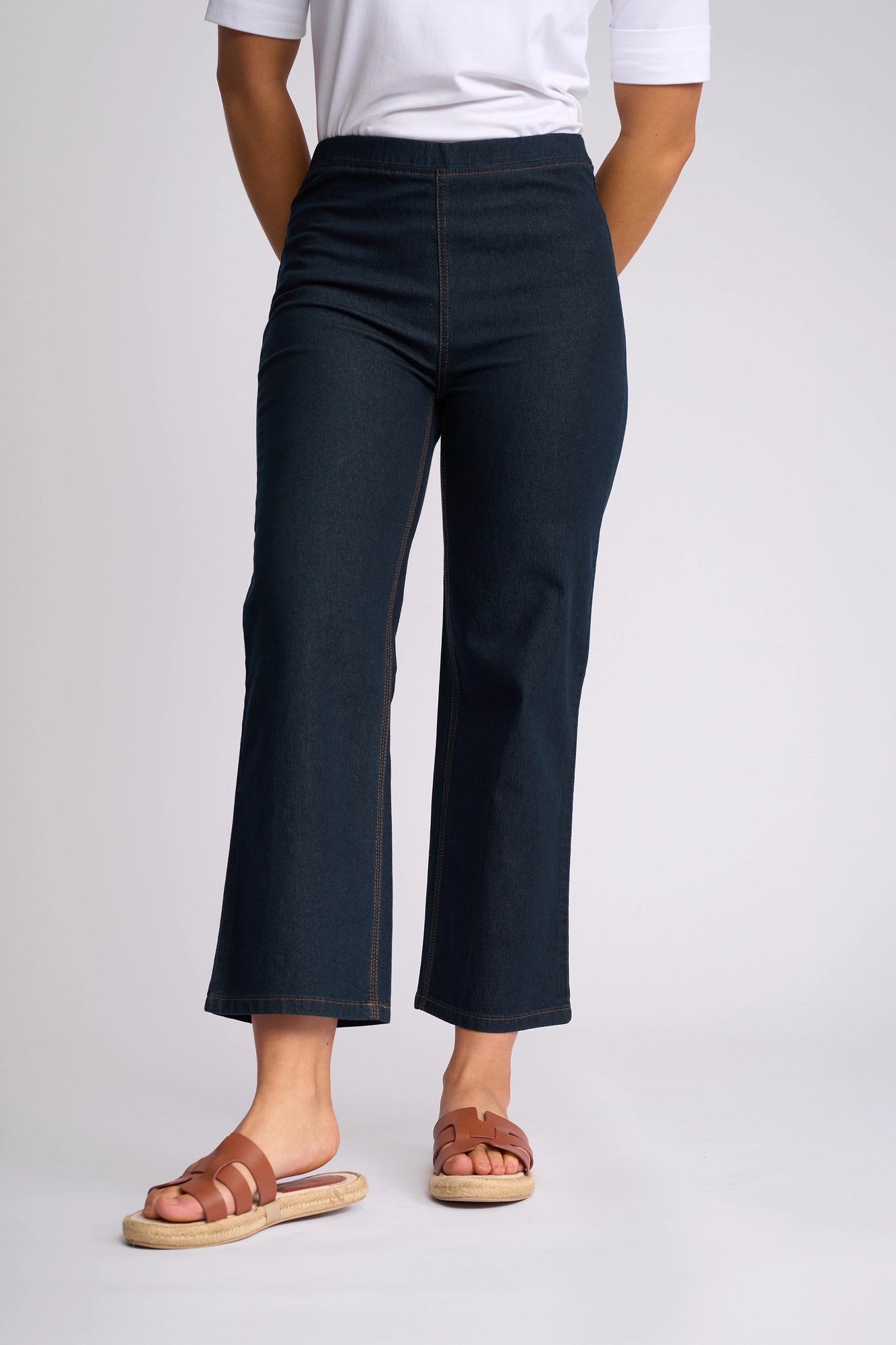A person is wearing the Vassalli Seattle Pull On - Indigo Denim pants, paired with a white top and brown sandals, standing against a plain light background. Only the lower half of their body is visible.