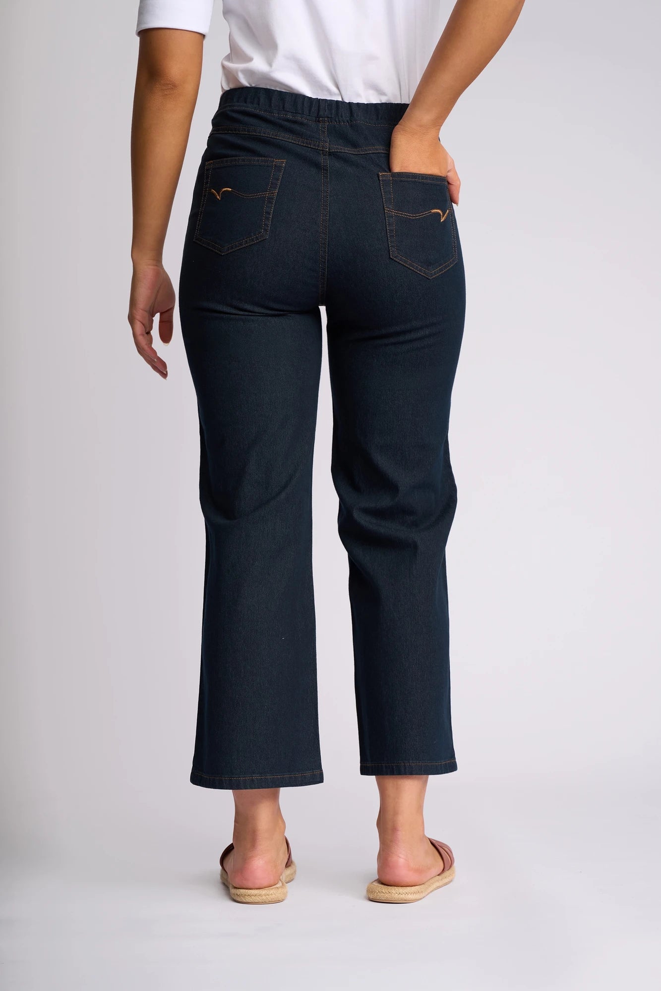 A person is shown from behind, standing with one hand in a back pocket and wearing the Vassalli Seattle Pull On - Indigo Denim jeans. The pull-on jeans feature flared legs and two back pockets, paired with a white shirt and sandals.