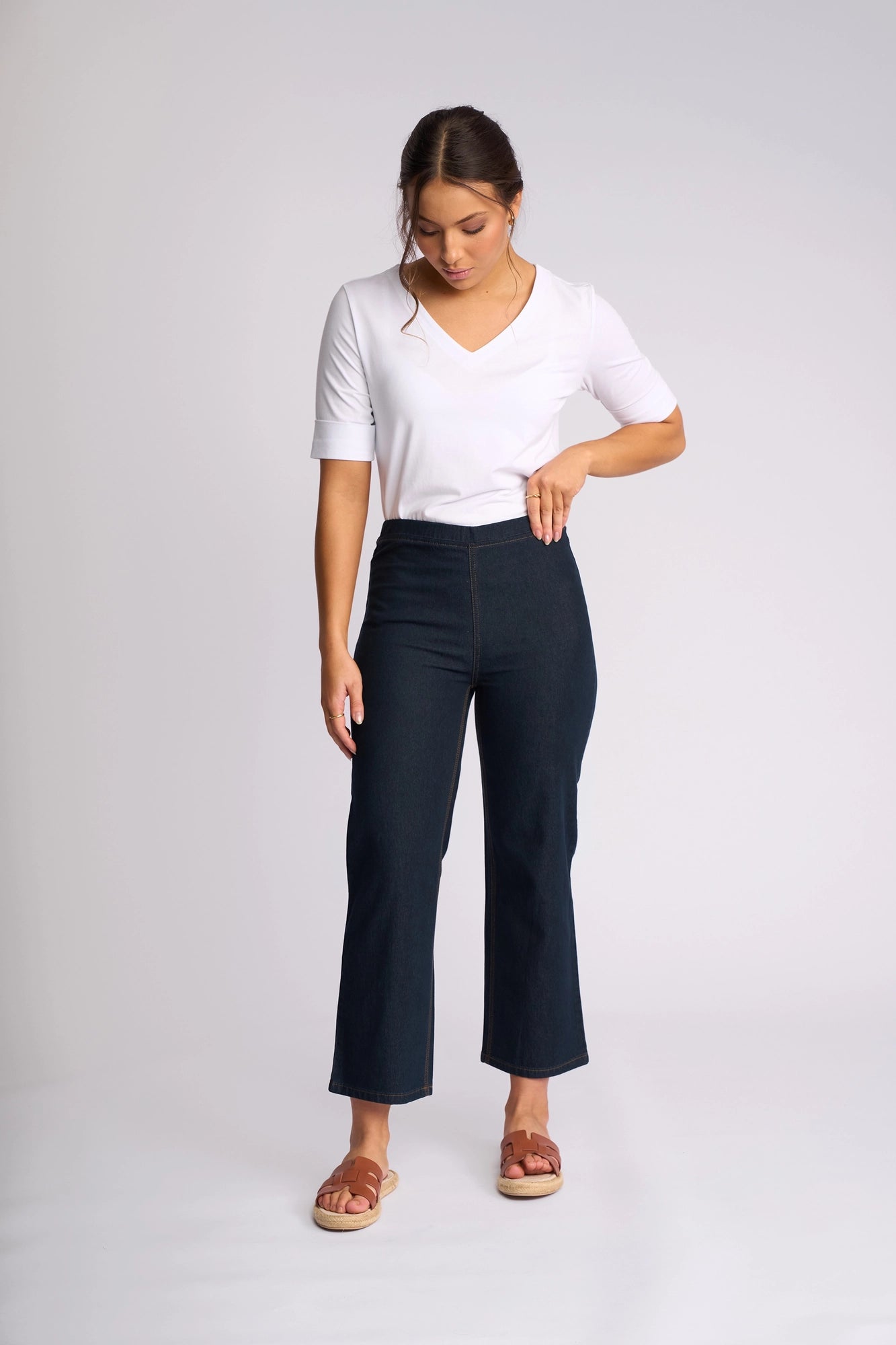 A woman wearing a white V-neck top, the Vassalli Seattle Pull On - Indigo Denim straight-leg jeans, and brown sandals stands against a plain light background, looking down with one hand on her hip.