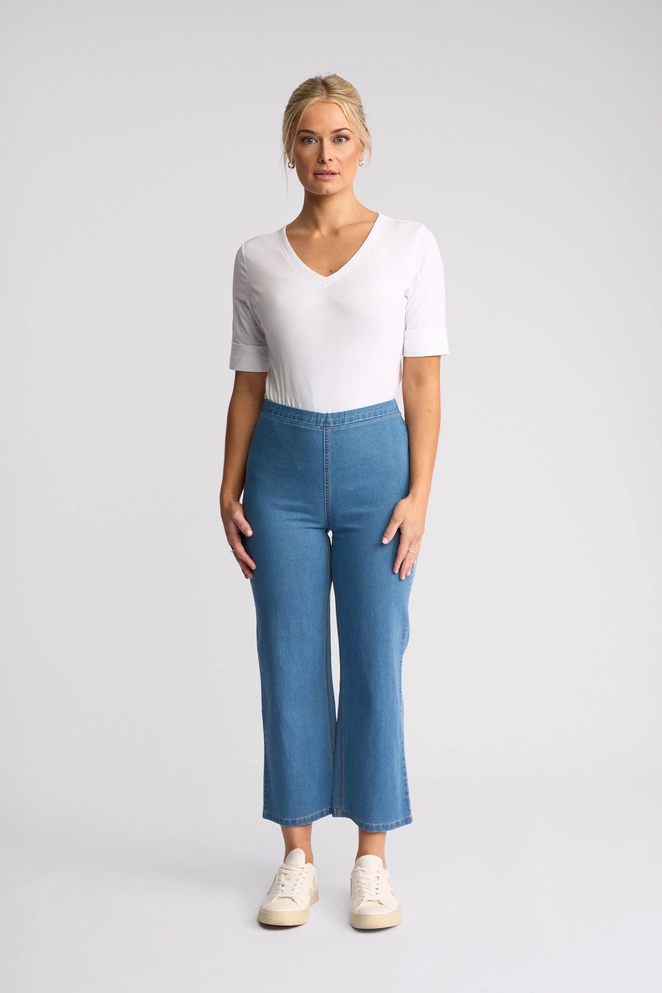 A woman stands against a white background, looking straight at the camera with a neutral expression. She is wearing a white V-neck shirt, white sneakers, and the Vassalli Seattle Pull On - Light Denim jeans: high-waisted, straight-leg jeans crafted from lightweight stretch denim.