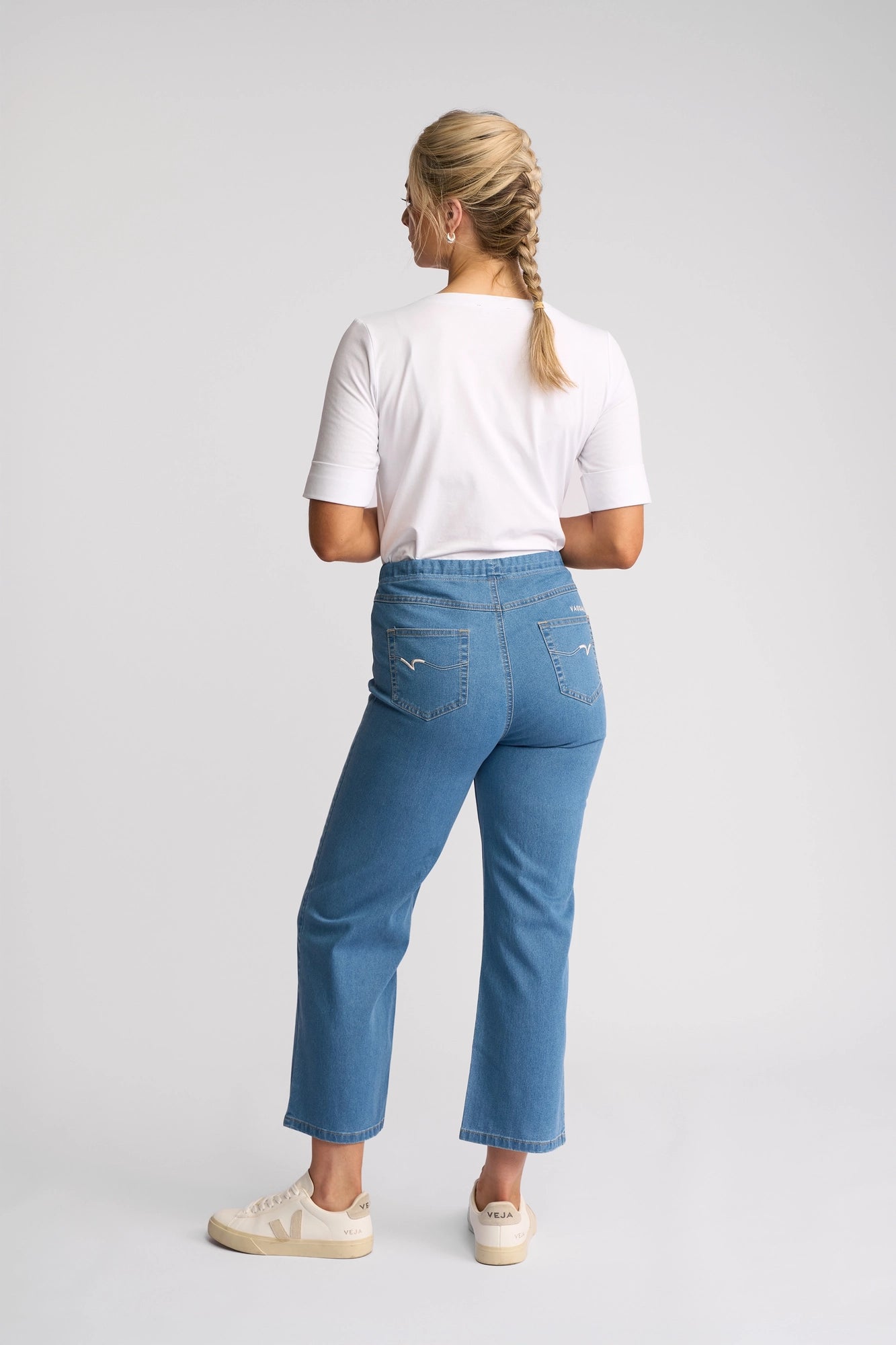 A woman with a blonde braid, dressed in a white shirt and Vassalli Seattle Pull On - Light Denim jeans, paired with beige sneakers, stands facing away against a plain white background.