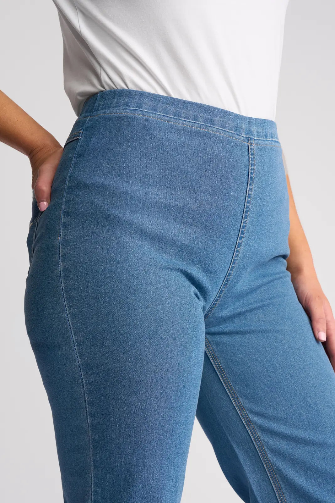 A person wearing Vassalli Seattle Pull On - Light Denim jeans, crafted from lightweight stretch denim, paired with a white top. One hand is tucked in their pocket as the image focuses on the waist and upper legs, highlighting the straight-leg fit and texture of these high-waisted pull on jeans.