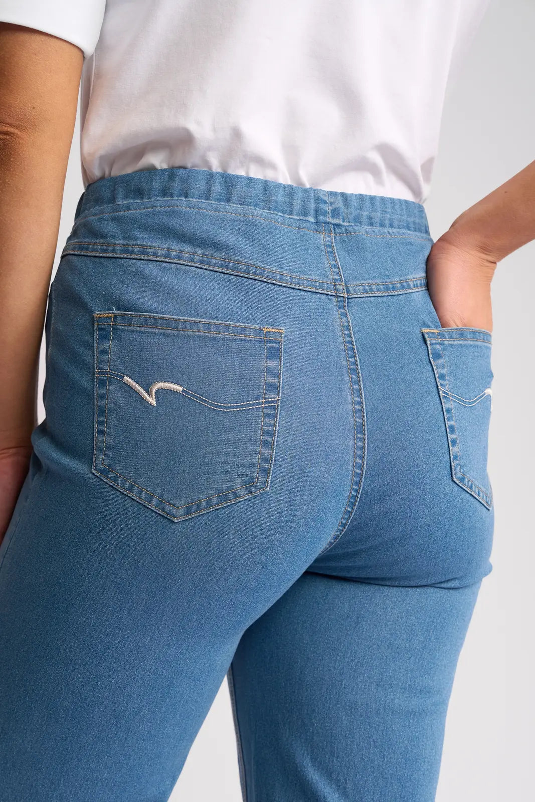 Close-up of a person wearing the Vassalli Seattle Pull On - Light Denim jeans and a white shirt. The focus is on the back pocket area of these lightweight stretch denim jeans, with one hand resting in the right back pocket.