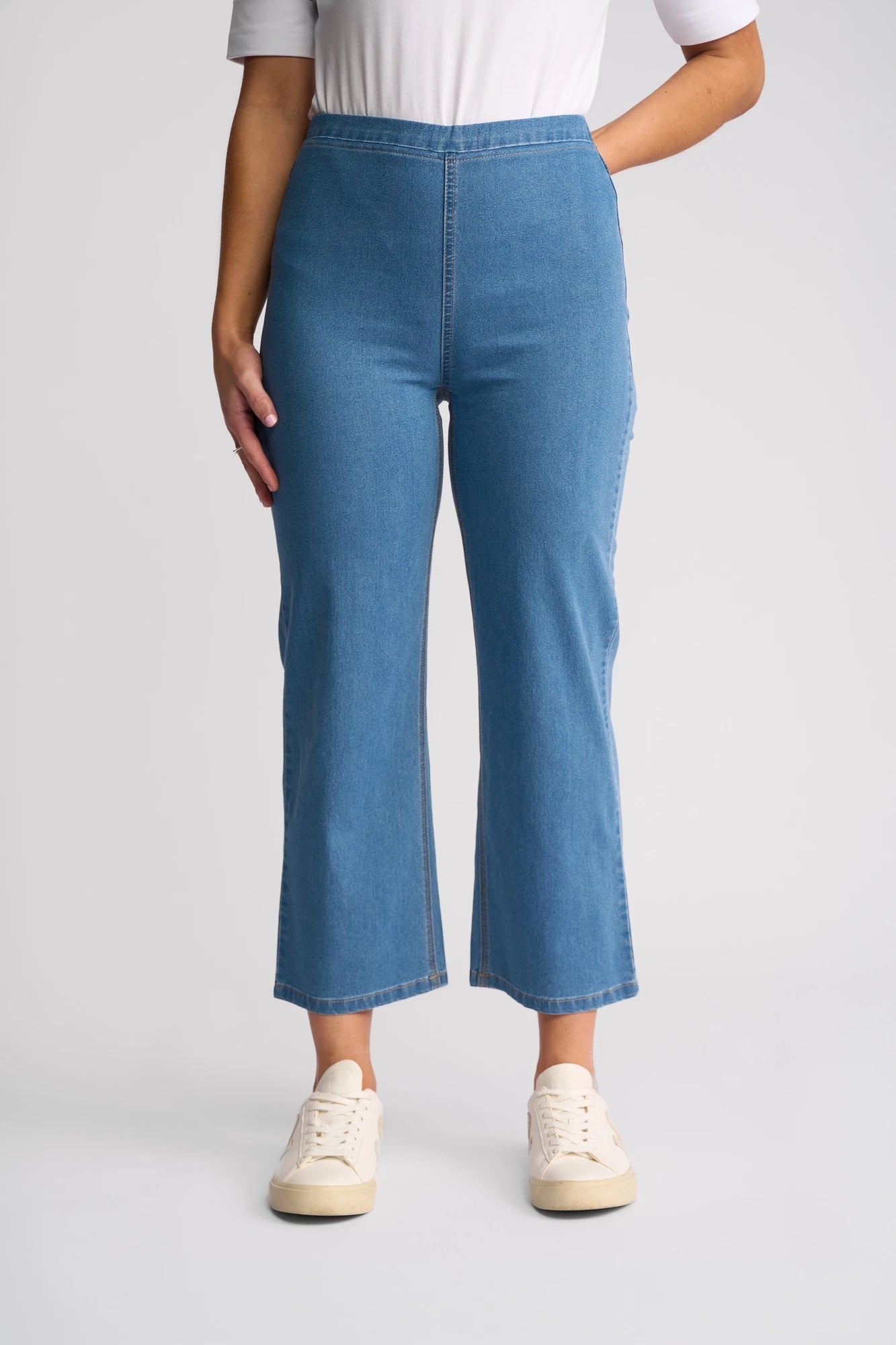 A person wearing a white short-sleeve shirt, white sneakers, and the Vassalli Seattle Pull On - Light Denim high-waisted straight-leg jeans crafted from lightweight stretch denim with a slight flare at the bottom, standing against a plain light background.