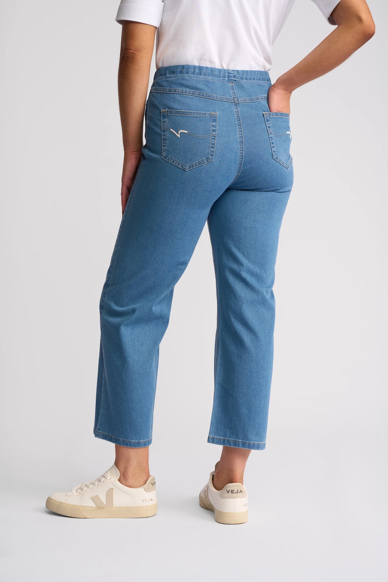 A person wearing a white t-shirt, Vassalli Seattle Pull On - Light Denim high-waisted straight-leg jeans crafted from lightweight stretch denim, and cream-colored Veja sneakers stands with one hand on their hip, facing away from the camera against a plain background.