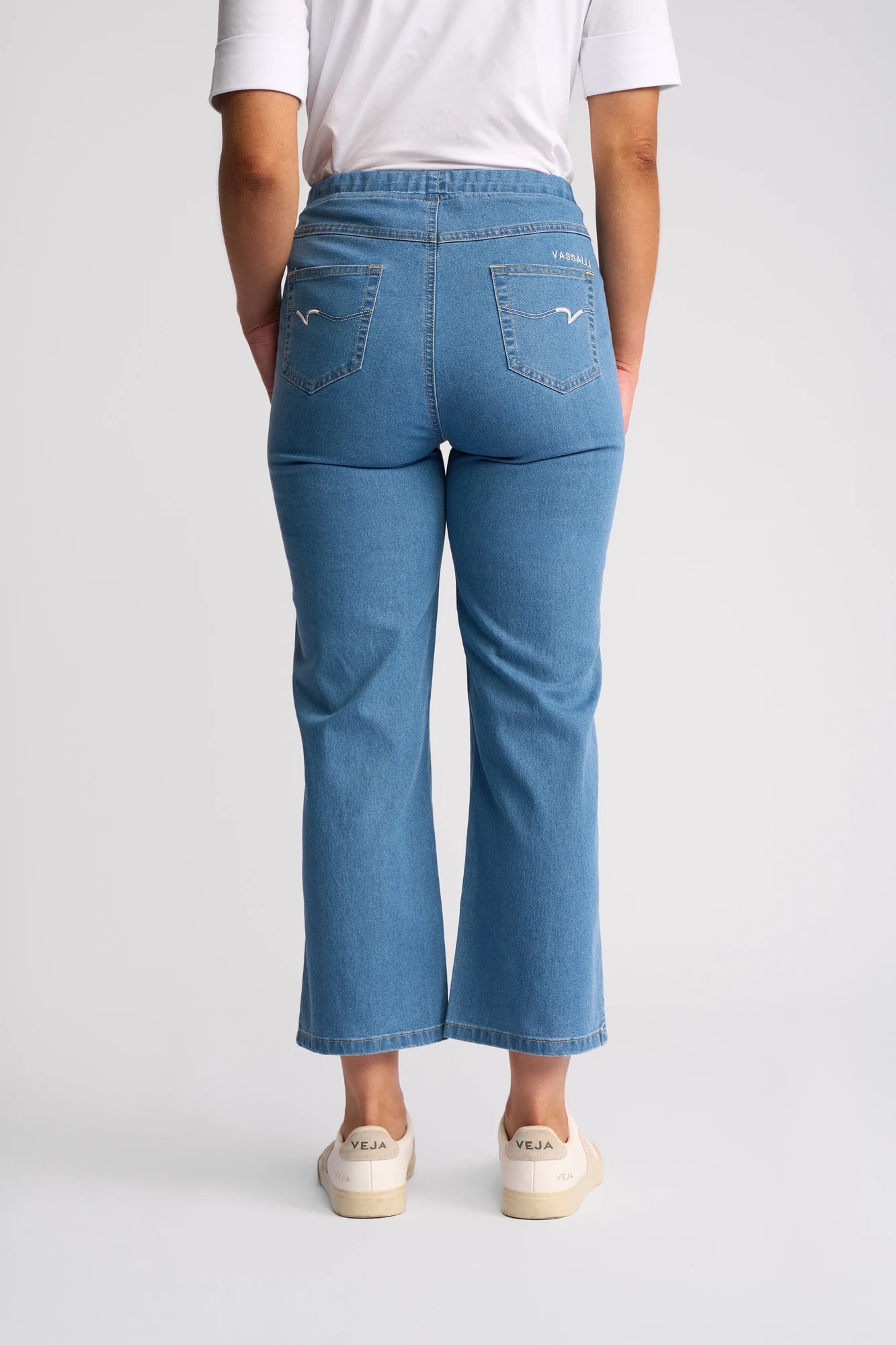 A person is shown from the back wearing Vassalli’s Seattle Pull On - Light Denim jeans with a straight-leg fit and a white T-shirt. They have paired the outfit with beige Veja sneakers, set against a plain, light-colored background.