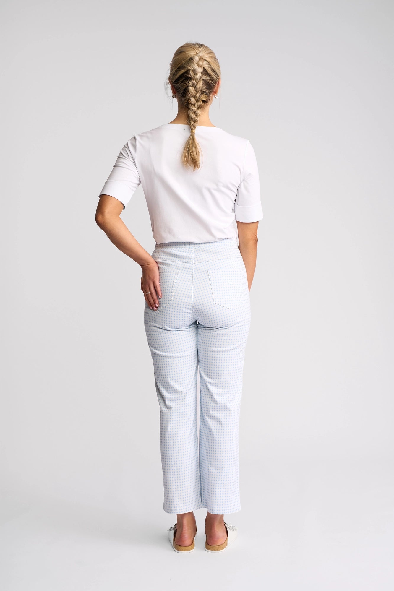 A woman with blonde braided hair stands facing away, wearing a white short-sleeve shirt, Vassalli Seattle Pull On pants in blue gingham stretch cotton, and cream-colored shoes. One hand rests on her lower back against a plain white background.