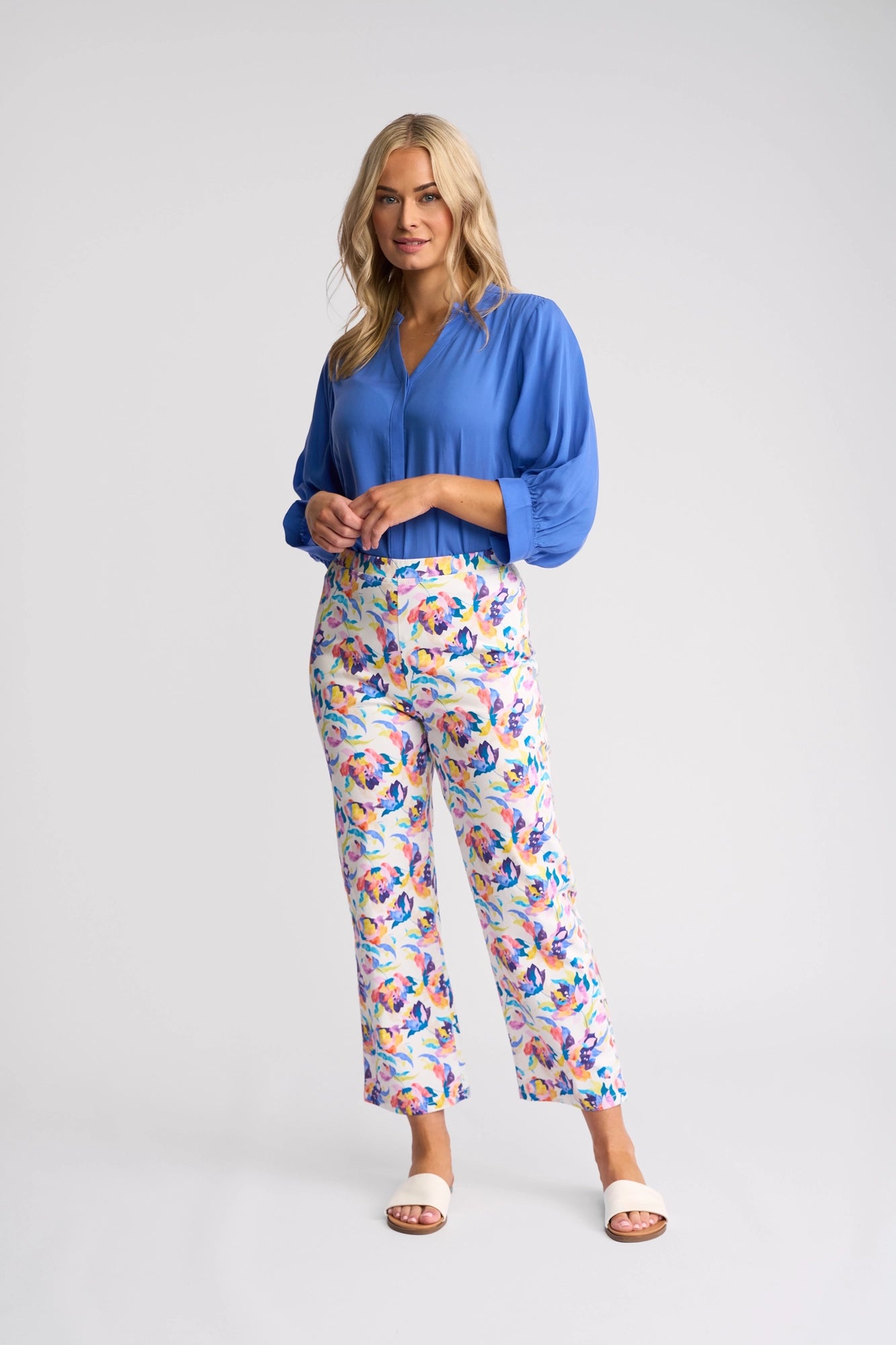 A woman with long blonde hair wears a blue blouse, the Vassalli Seattle Pull On - Demi colorful stretch cotton floral print pants, and white sandals, standing against a plain light background.